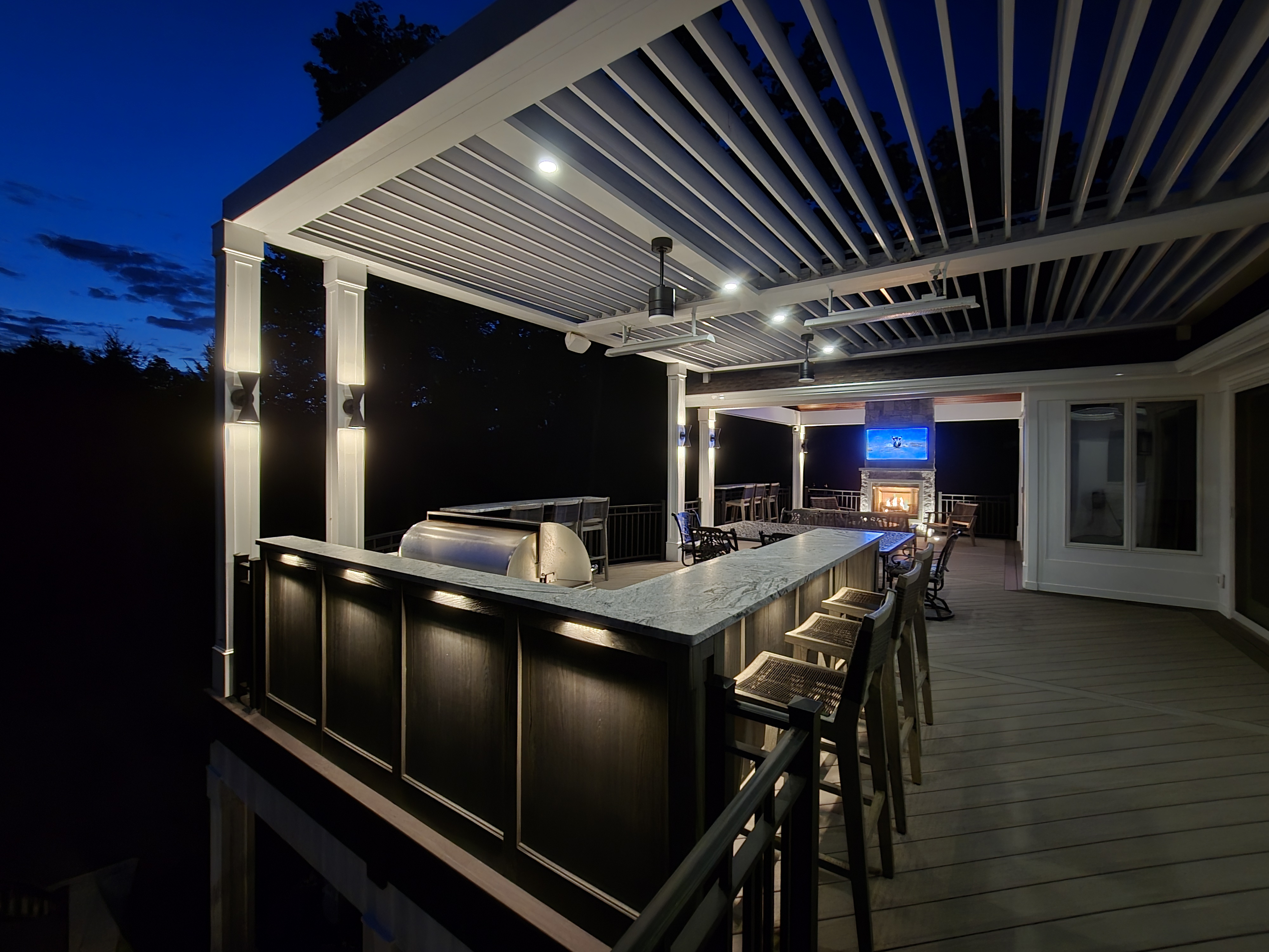 nighttime outdoor bar and lounge with white louvered roof, stone fireplace, and mounted TV