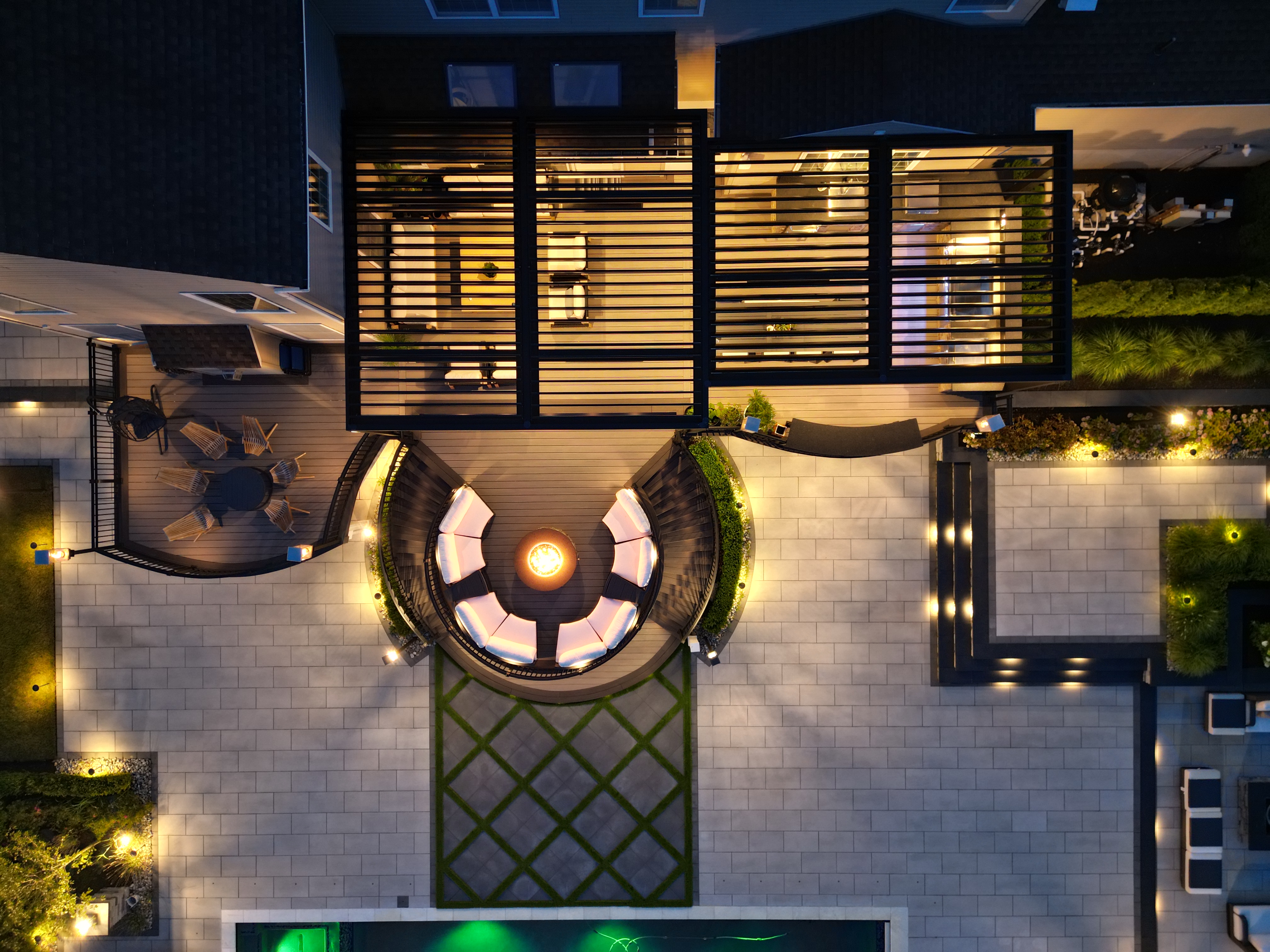 aerial view of luxury deck with dual louvered pergolas, circular fire pit lounge, outdoor kitchen, and tiered patio with poolside lighting