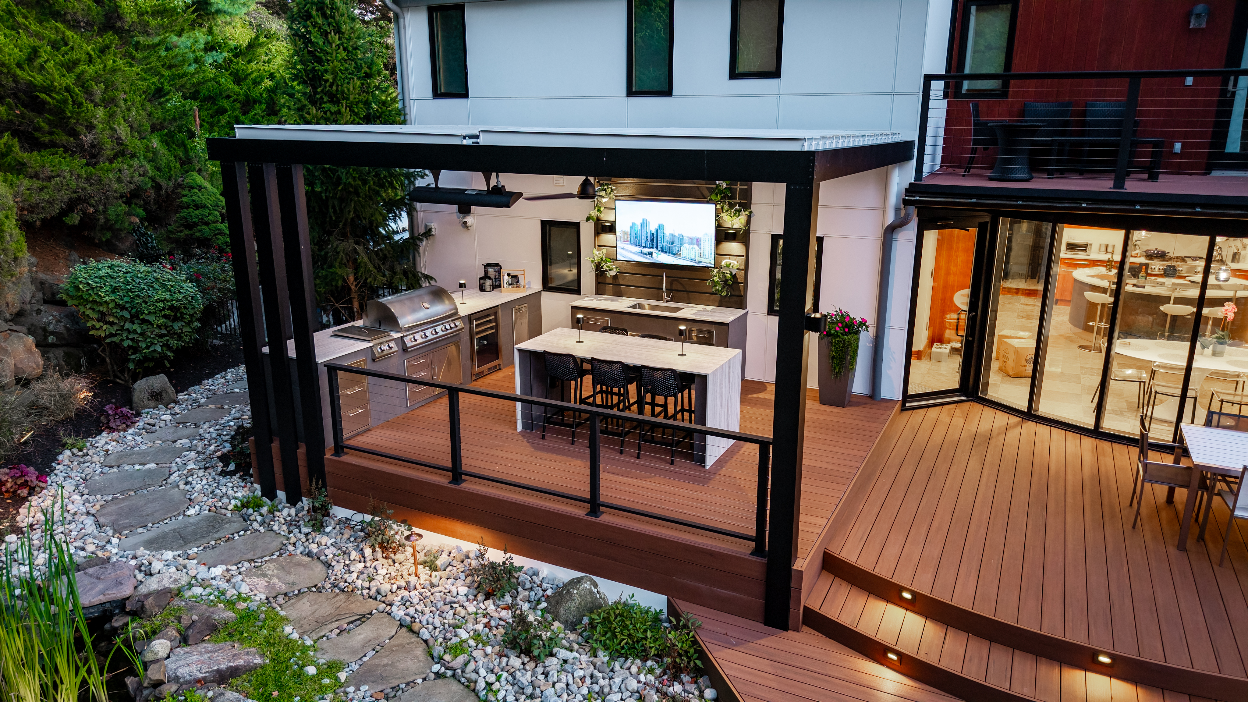 raised deck with outdoor kitchen, island seating, and louvered pergola beside a stone garden path
