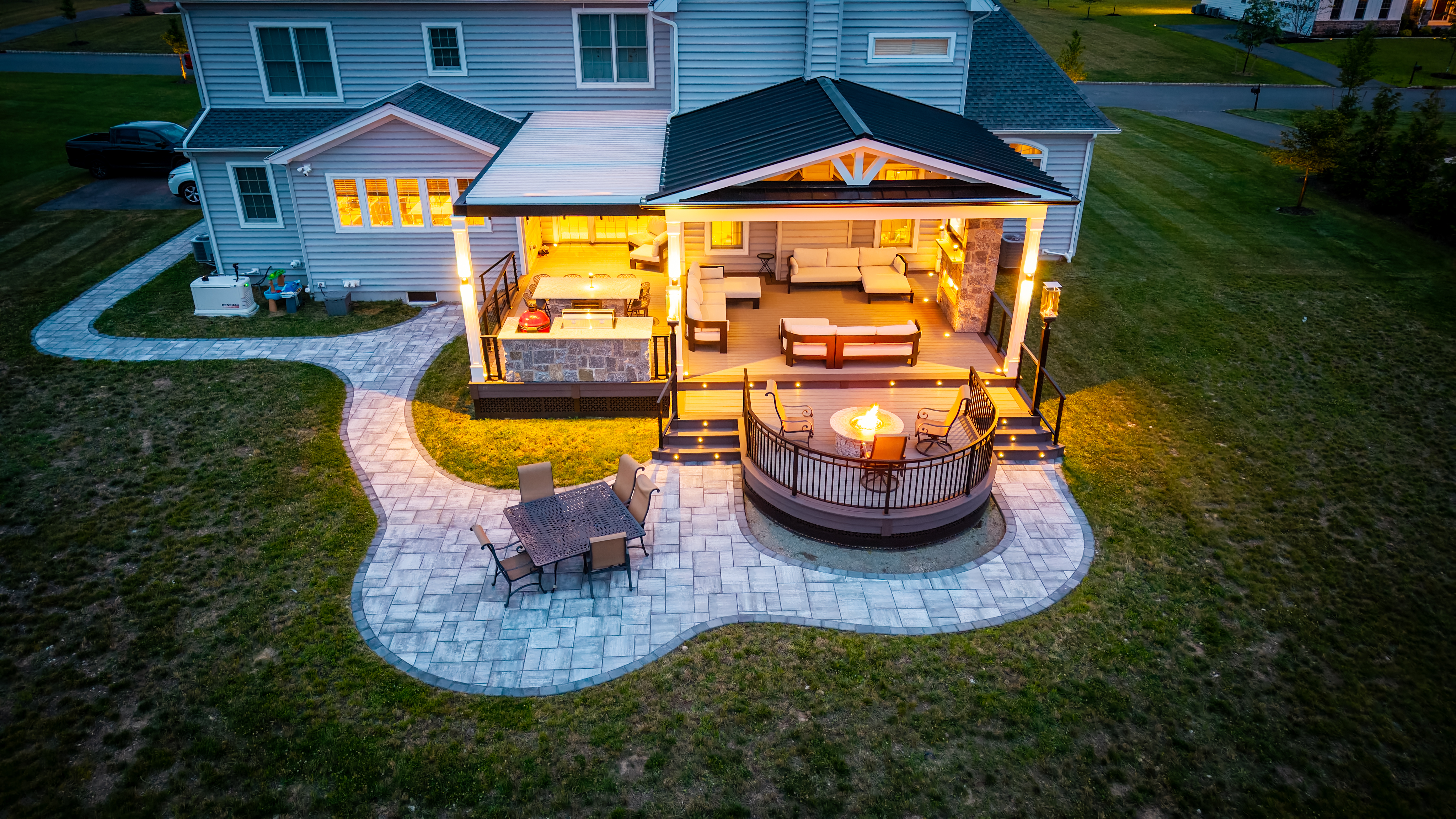 covered backyard deck with louvered roof, outdoor kitchen, lounge seating, curved fire pit area, and paver patio with step lighting
