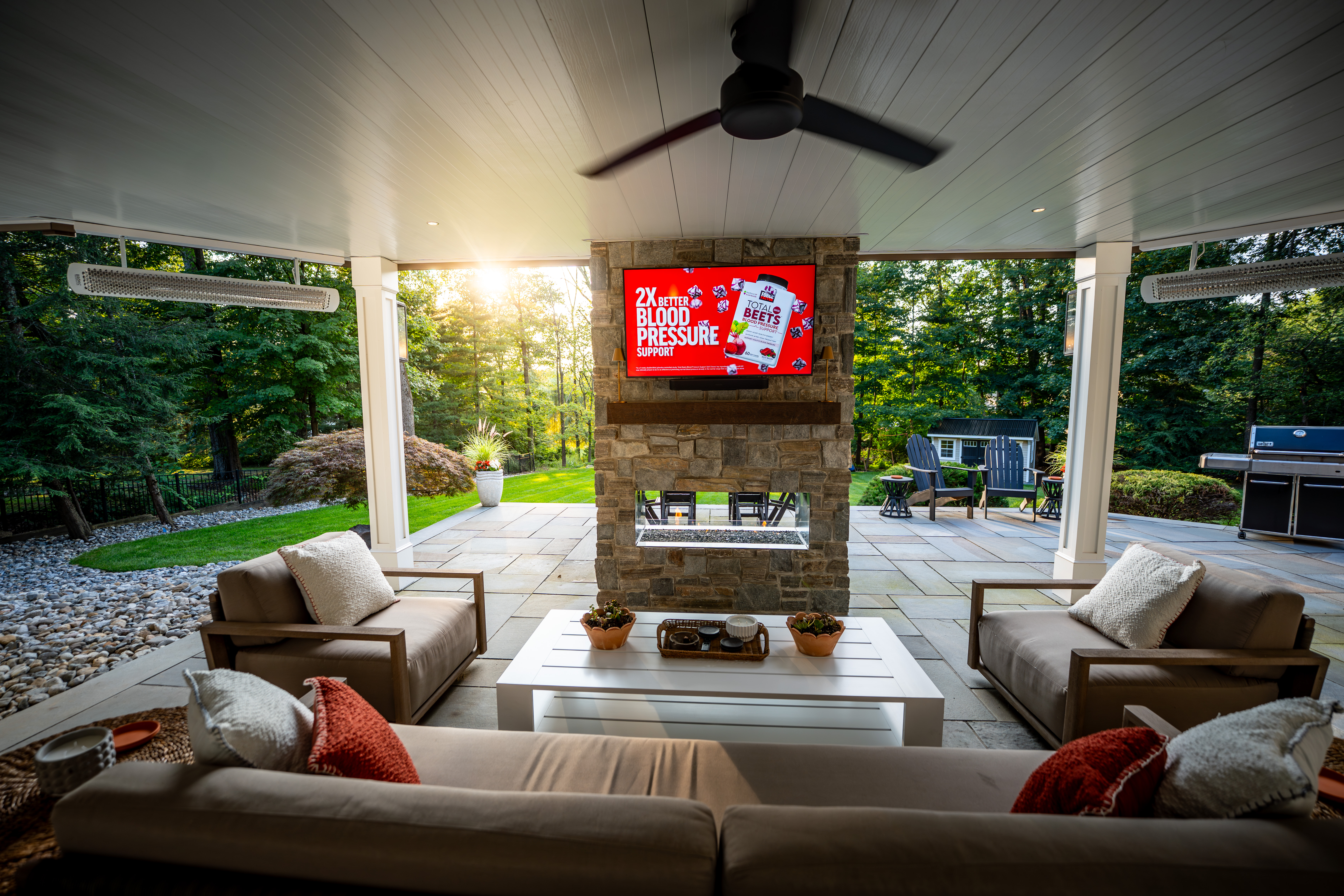 finished under-deck patio with stone fireplace, mounted TV, and cushioned seating surrounded by greenery