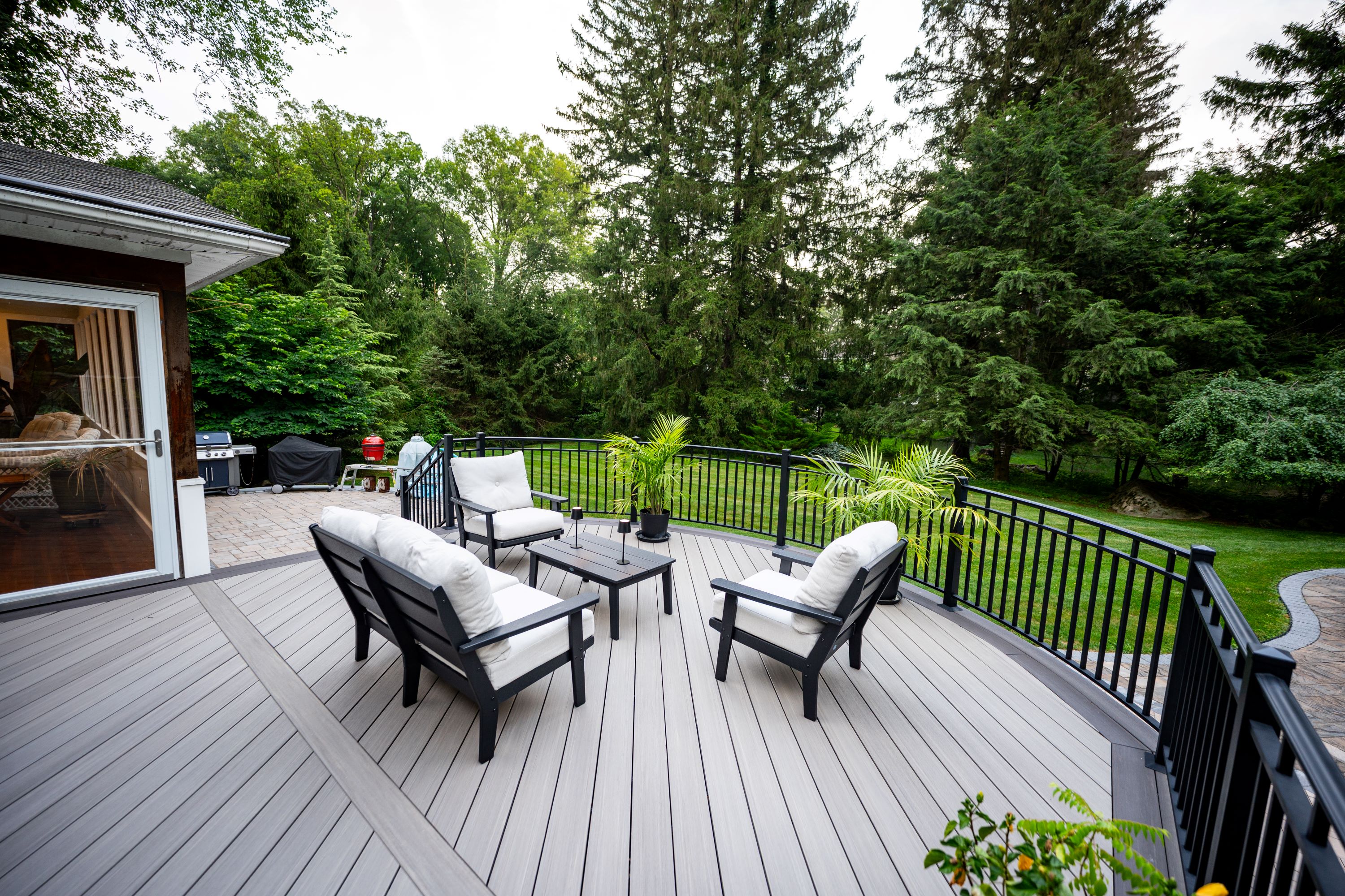 curved deck with black railing and outdoor seating overlooking large wooded backyard and paver patio
