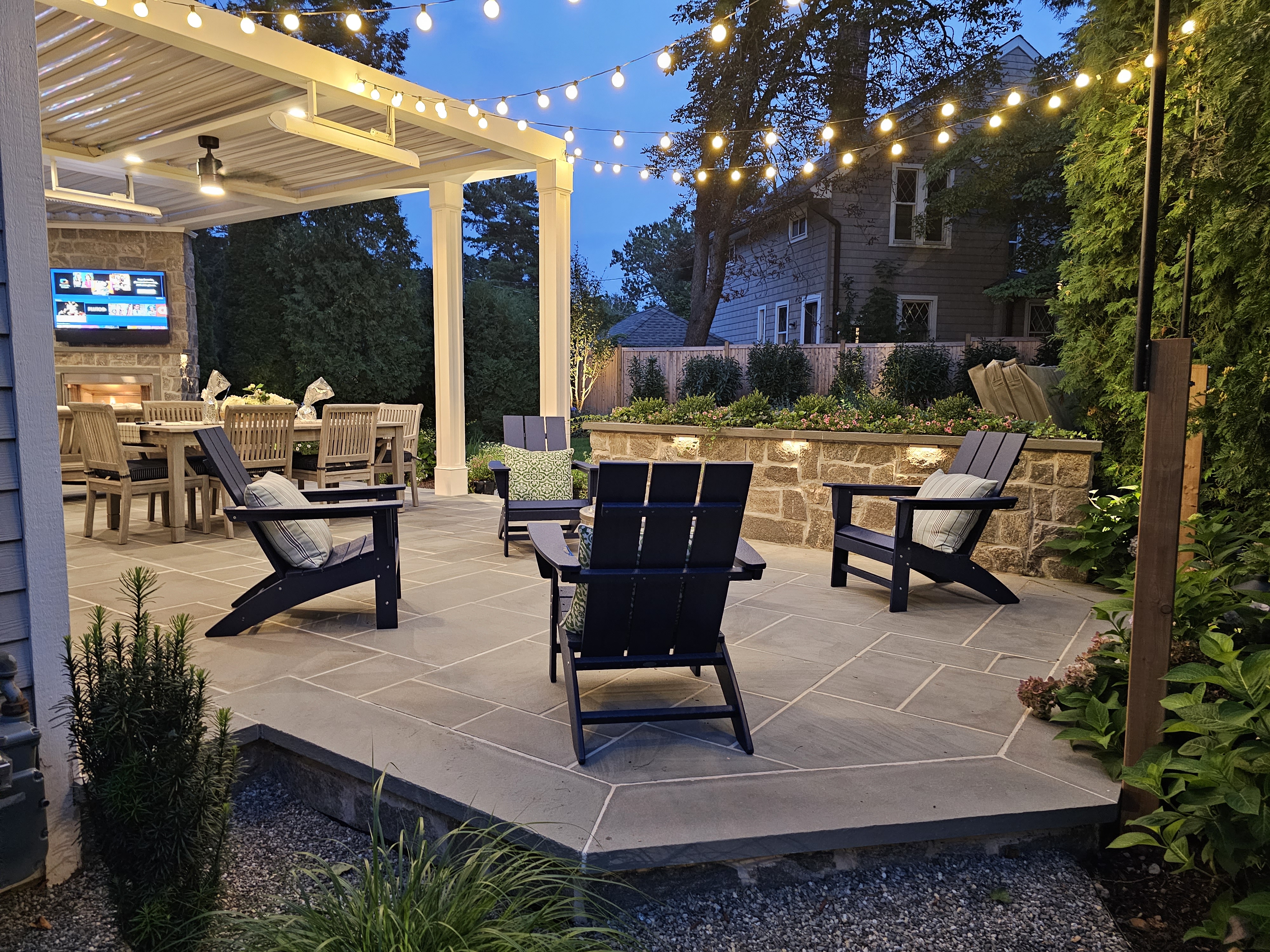 luxury paver patio with louvered roof, built-in outdoor kitchen, fire pit seating area, string lights, and stone retaining wall with accent lighting