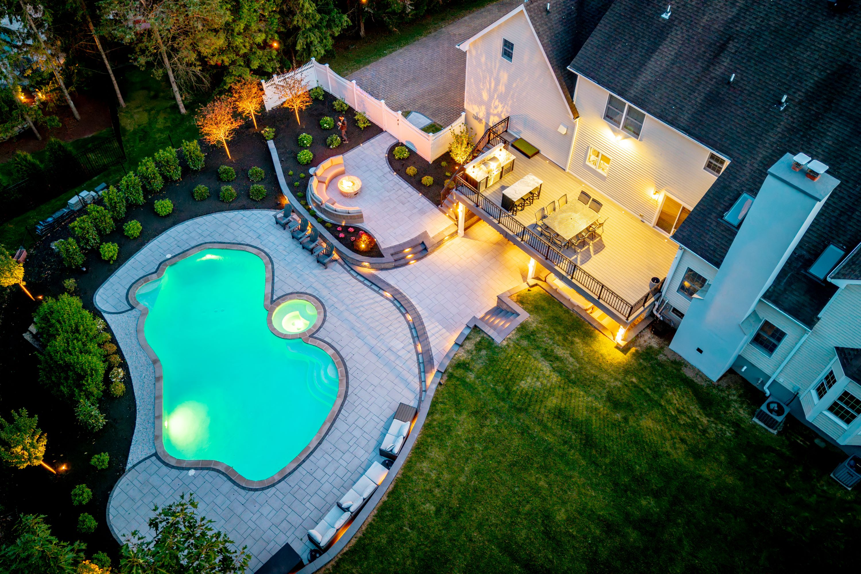 aerial view of elevated deck with outdoor kitchen and dining area, paver patio with fire pit seating, and freeform pool with spa