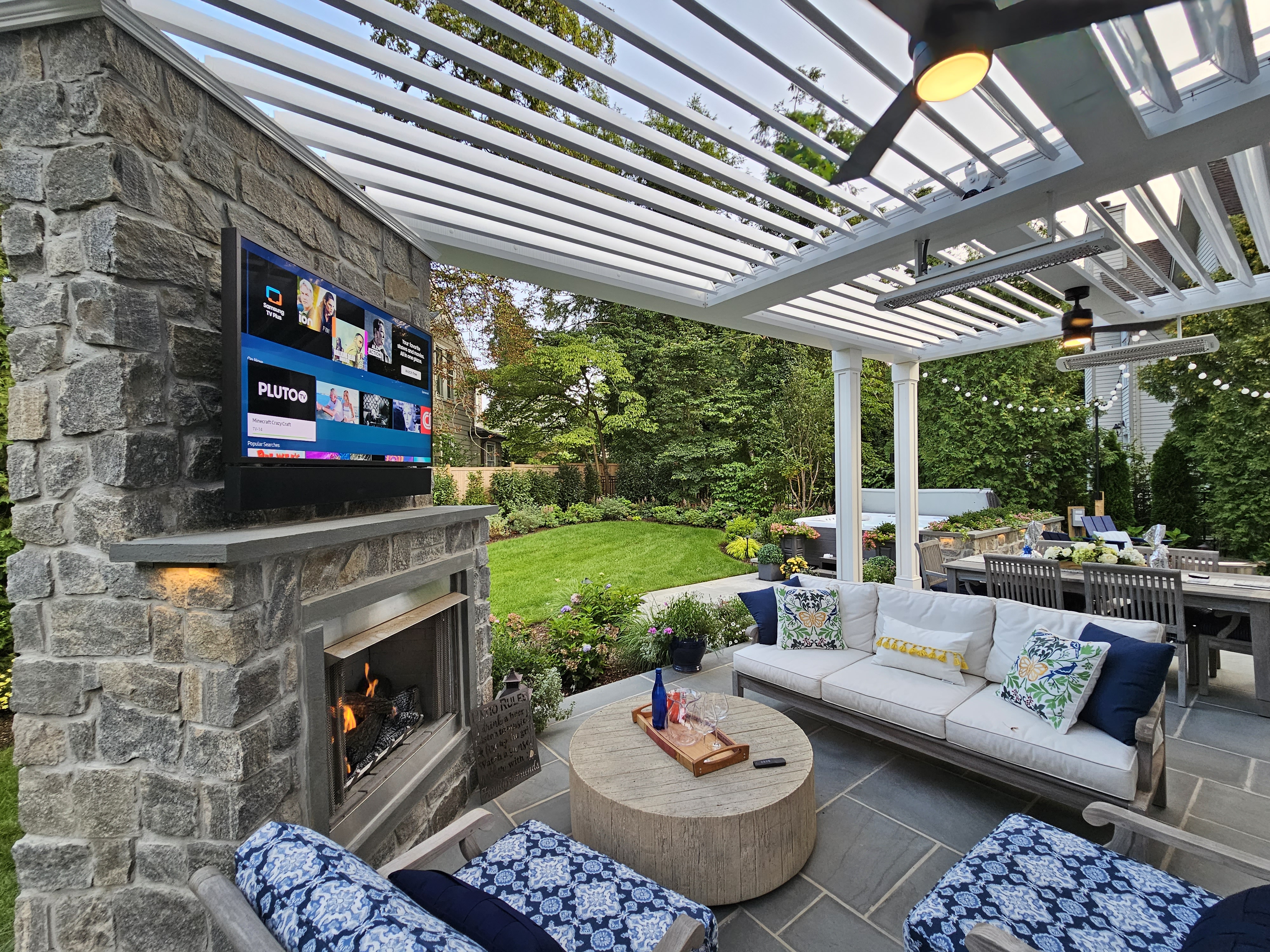 outdoor living room with stone fireplace, mounted TV, and white louvered roof over patio seating and dining areas