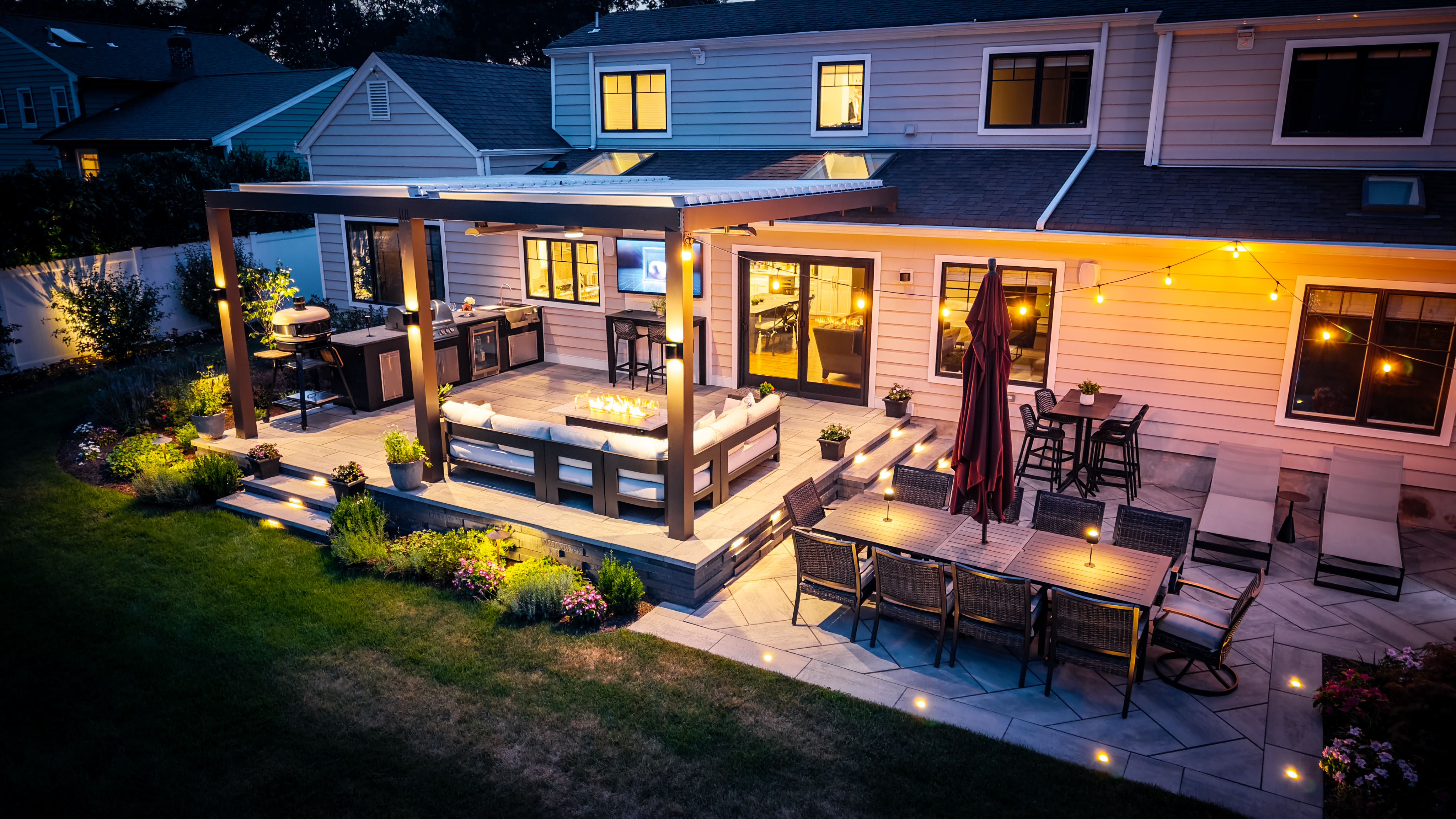 backyard outdoor living space with pergola covered kitchen, fire feature seating, and patio dining area at dusk
