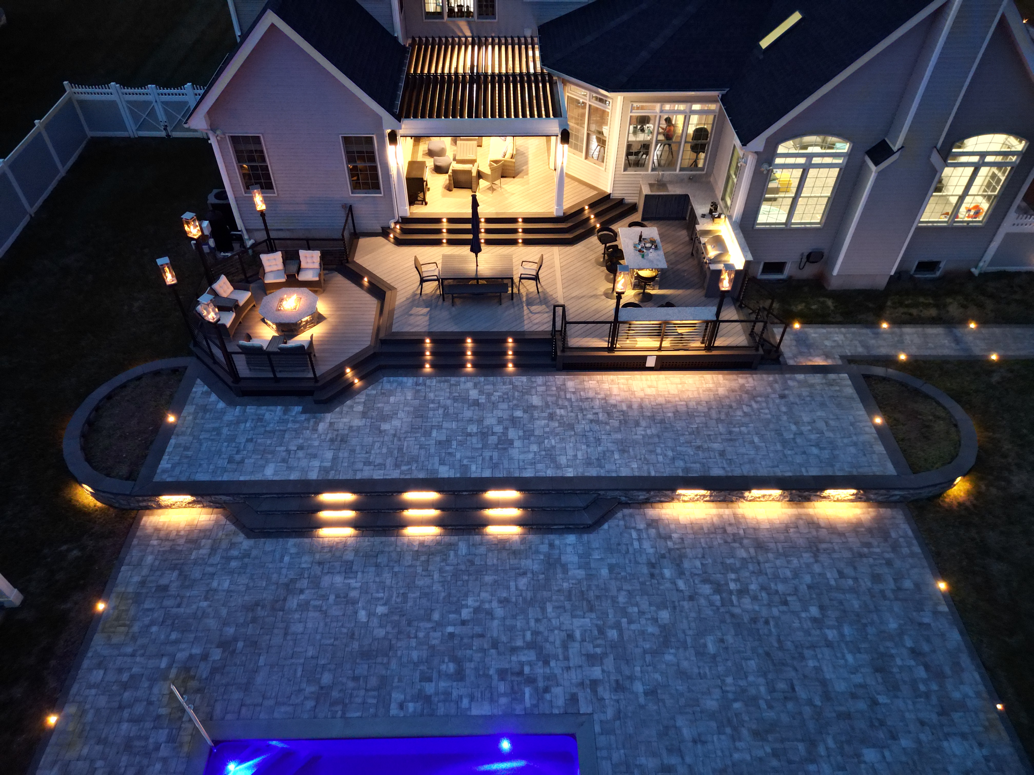 elevated deck with fire pit seating, dining area, and built-in kitchen overlooking a large stone patio with lighting and pool