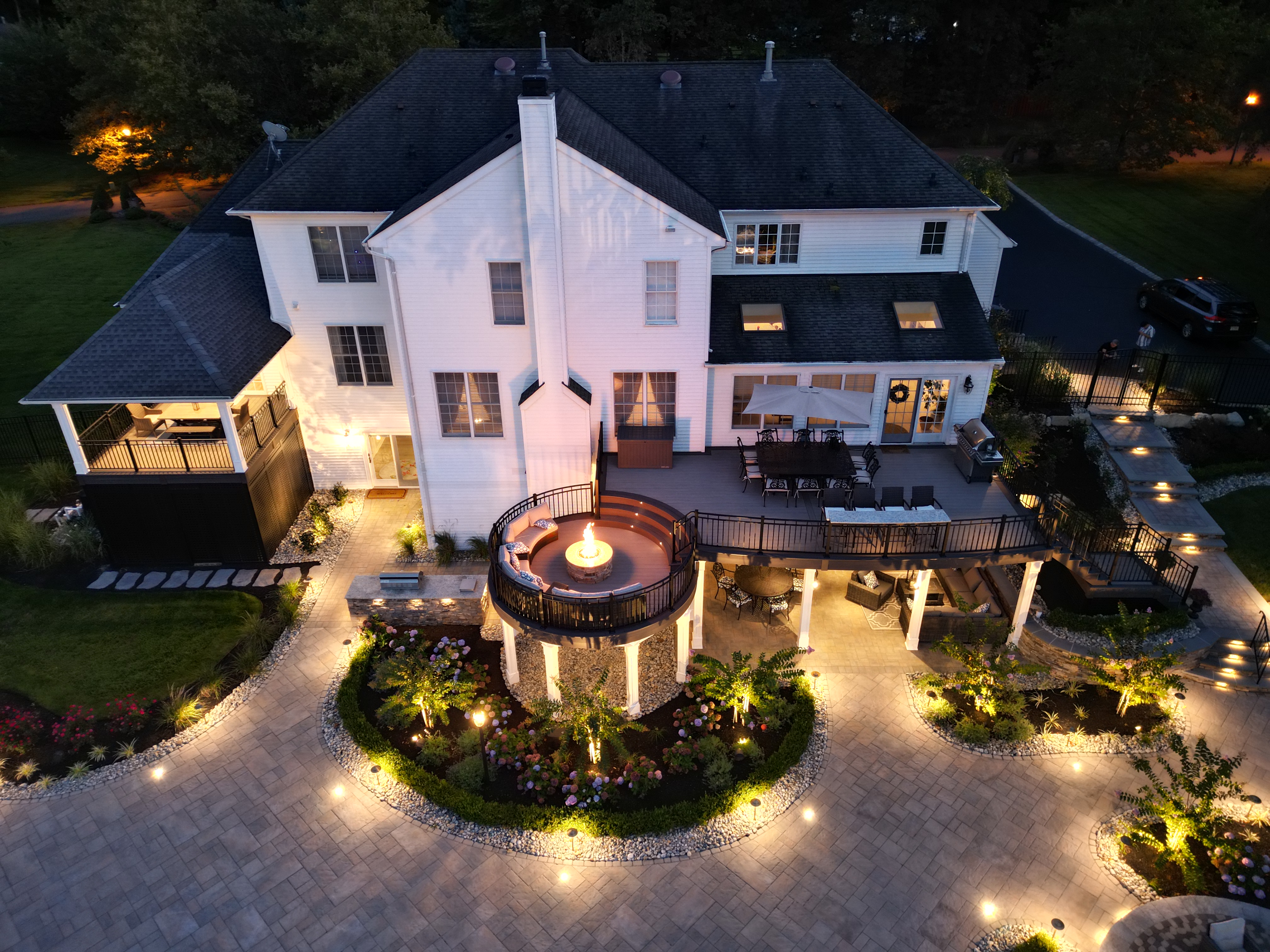 multi-level deck with dining and lounge area, elevated circular fire pit seating, and lower covered patio with outdoor furniture, surrounded by landscaped hardscaping and lighting