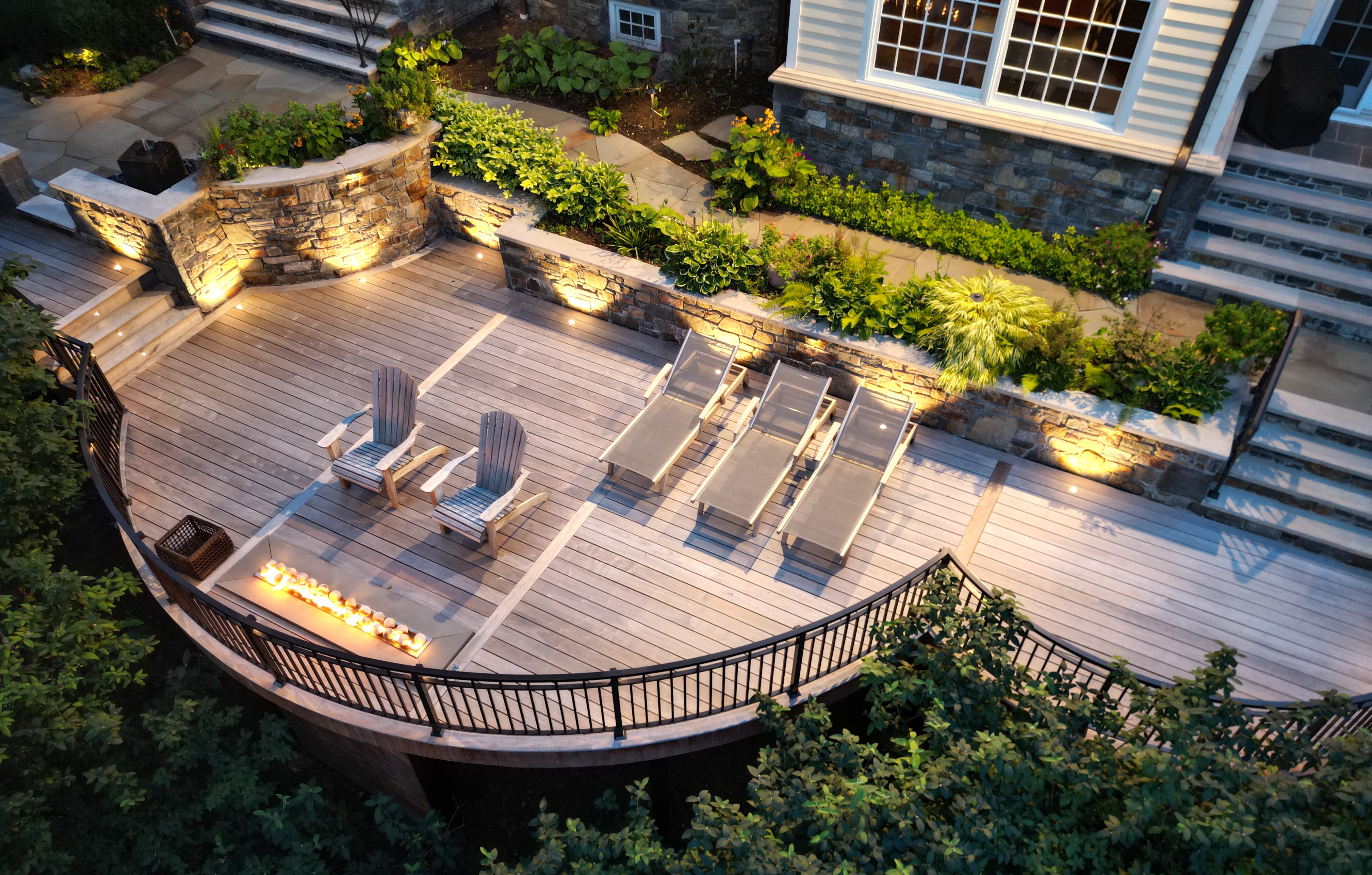 curved hardwood deck with integrated fire feature, built-in lighting, wood lounge chairs, sun loungers, and tiered stone retaining walls