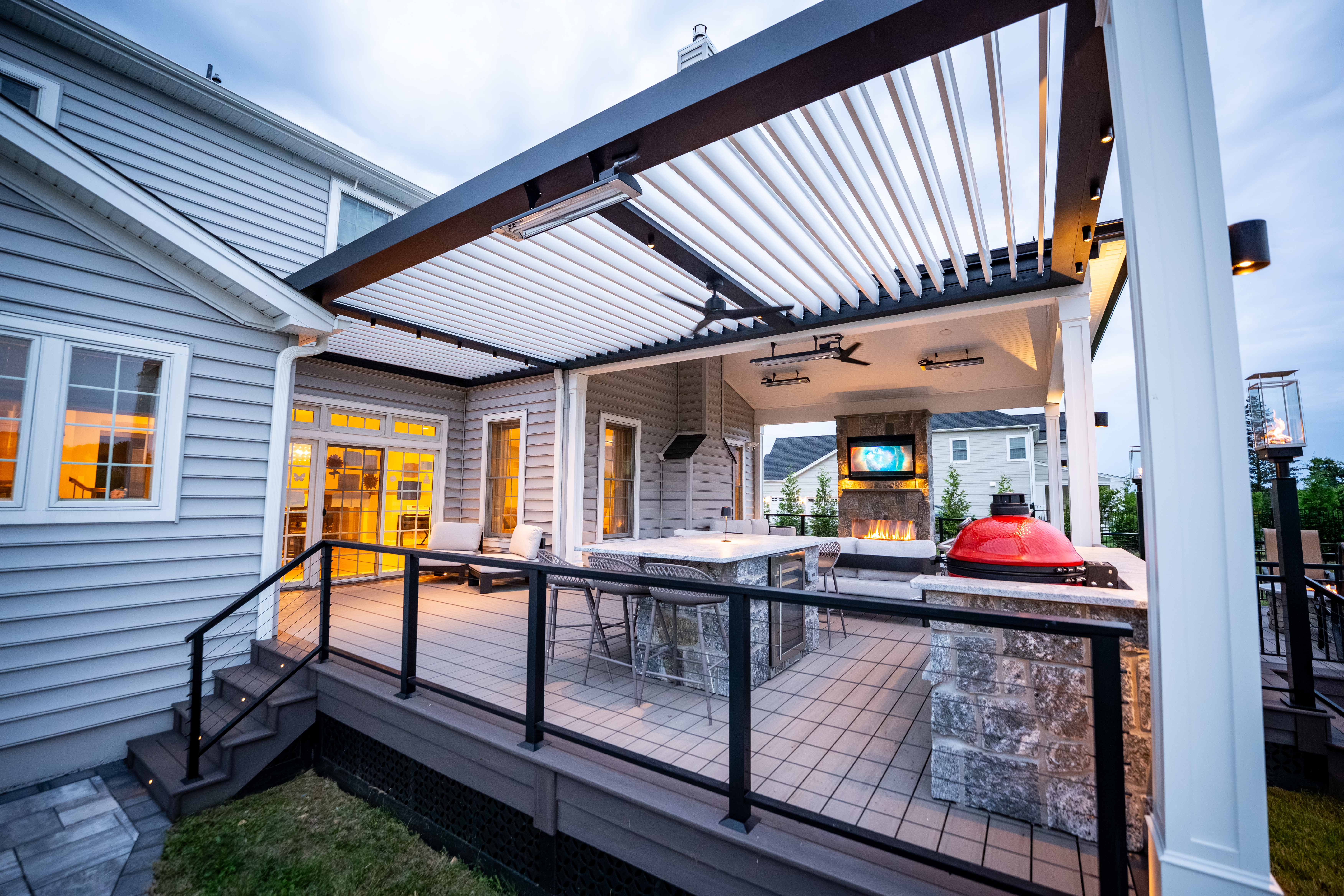 backyard deck with black and white louvered roof, outdoor kitchen with grill and pizza oven, stone bar seating, lounge area, and fireplace with TV