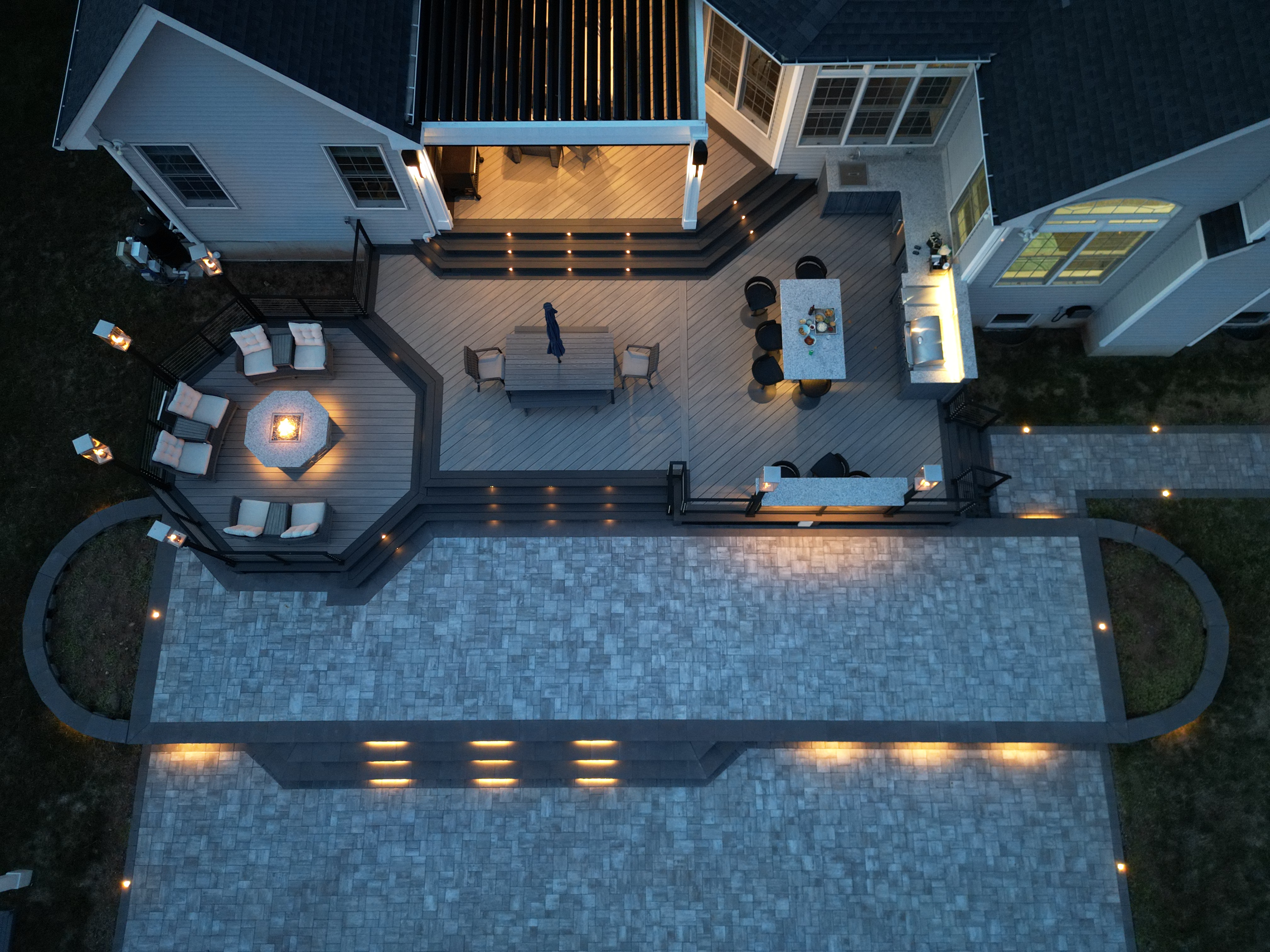 spacious backyard deck with patio lighting on steps, built-in grill station, and illuminated fire pit lounge area