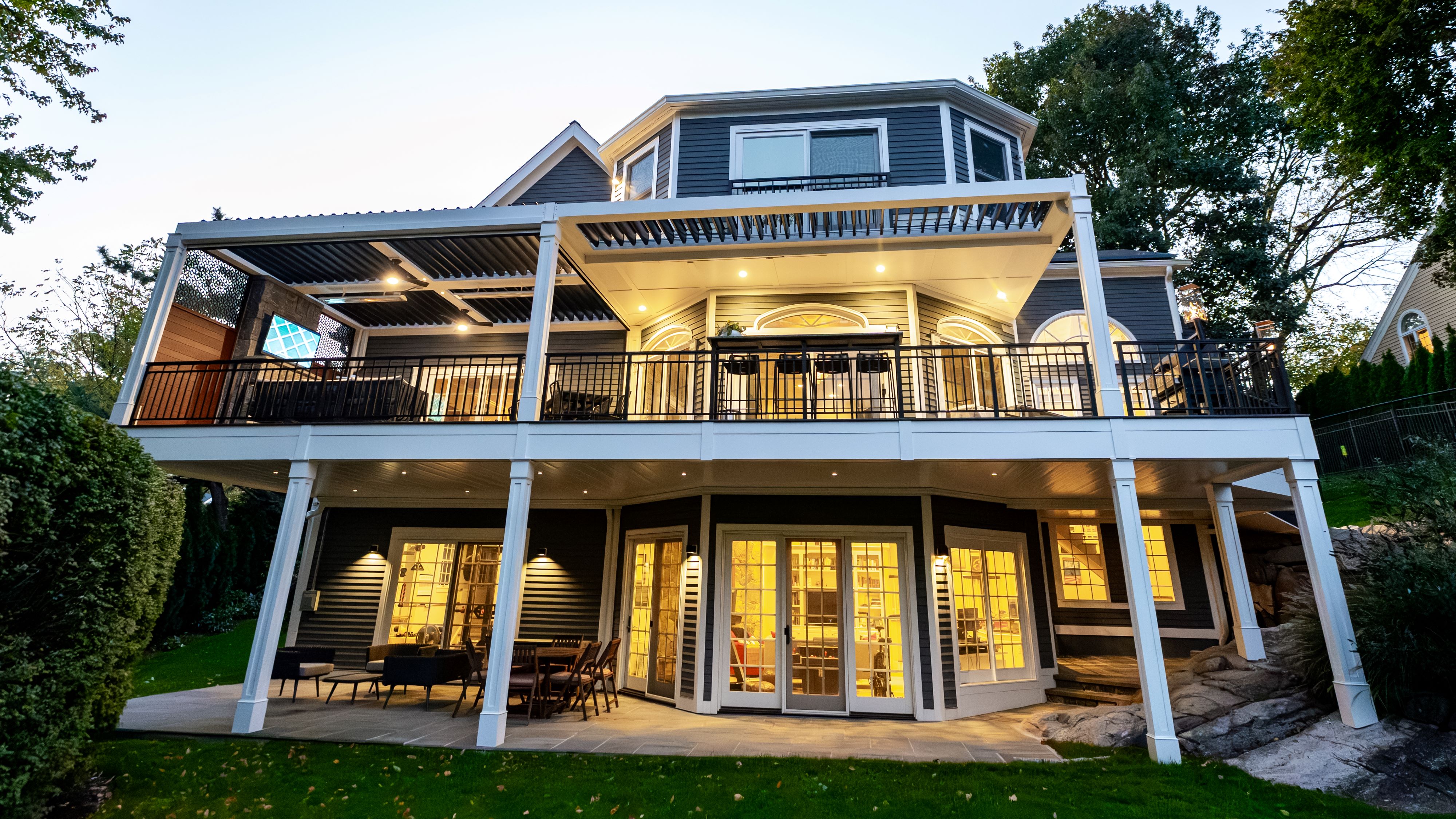 multi-level home exterior at dusk with finished under deck dining area, paneled ceiling, recessed lighting, white columns, French doors, and upper deck with louvered roof and black railing