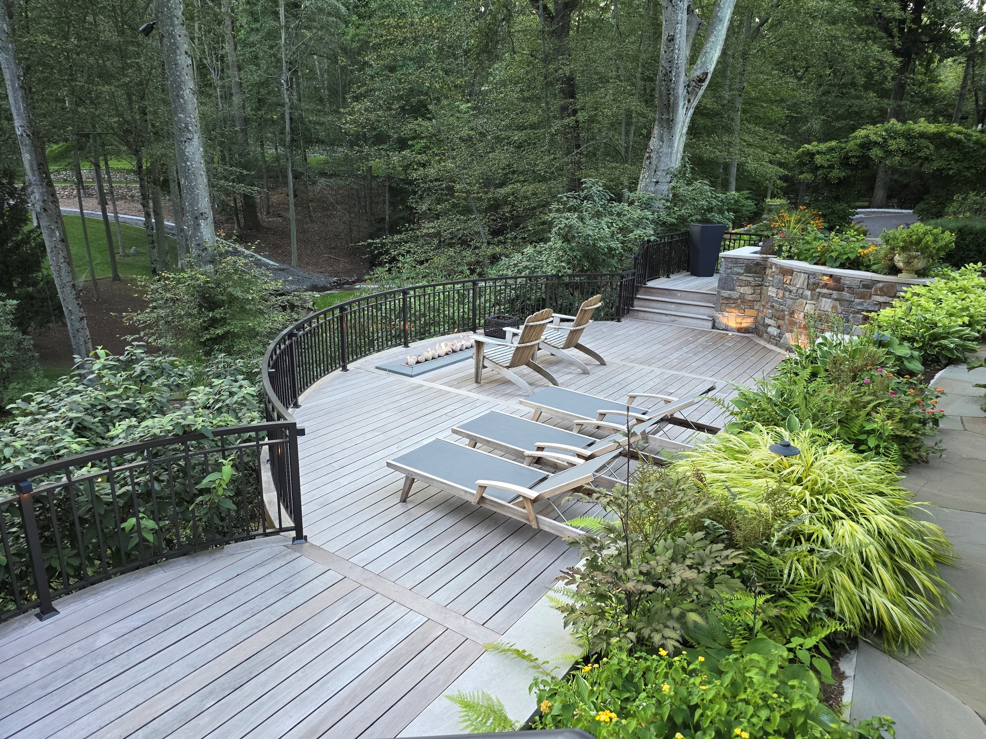 curved Ipe deck overlooking wooded backyard with lounge chairs, linear fire feature, black railing, and surrounding plantings