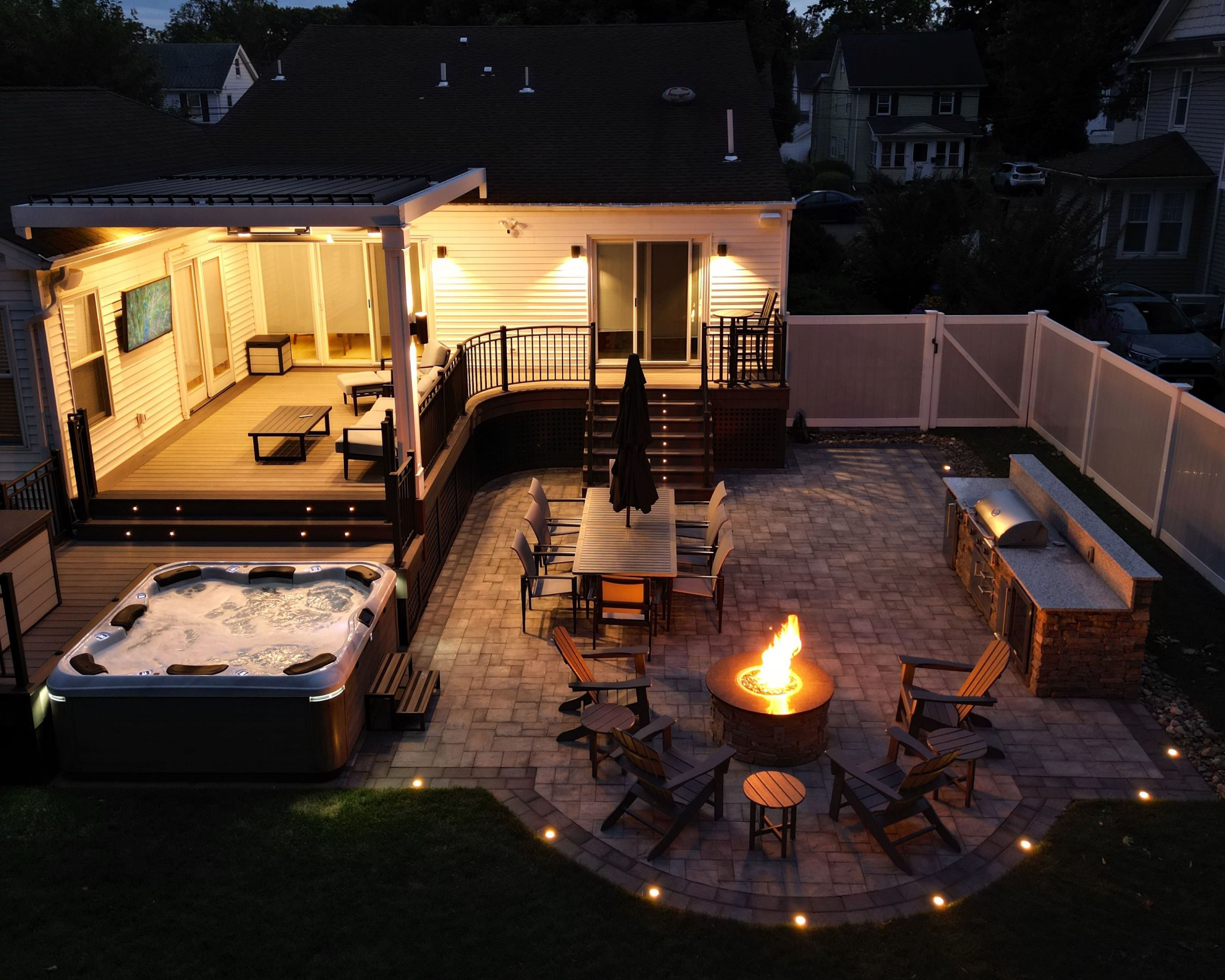multi-zone backyard outdoor living space with covered deck, hot tub, outdoor kitchen, dining area, and fire pit seating at dusk