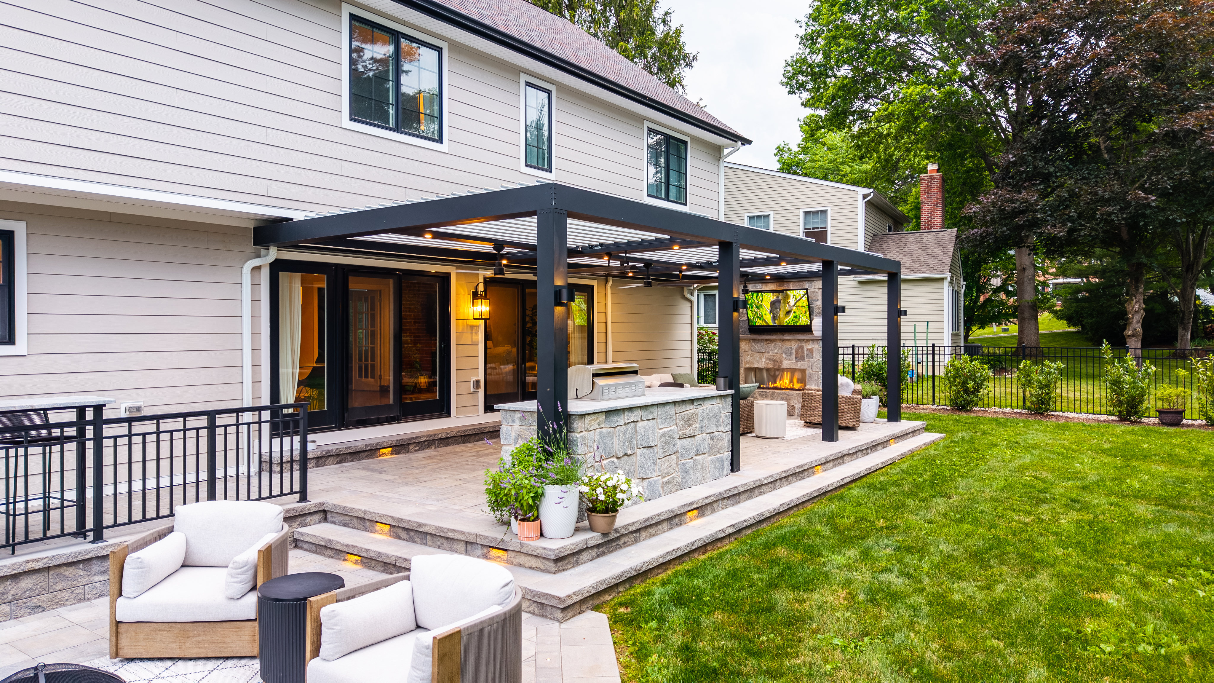 compact backyard patio with louvered roof pergola, stone outdoor kitchen, linear gas fireplace, and tiered paver steps with integrated lighting