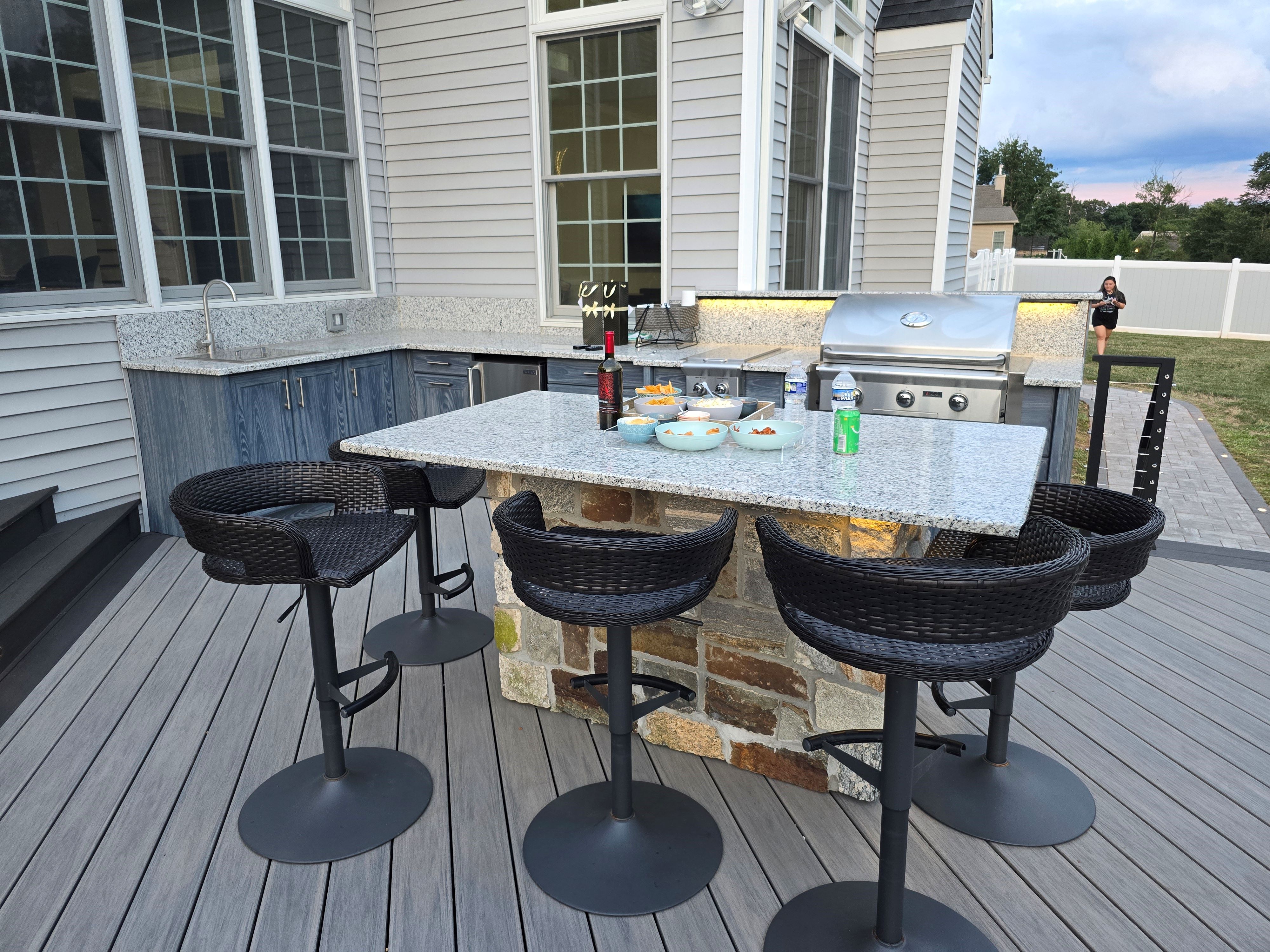 outdoor kitchen on a deck with granite island seating, built-in grill, sink, refrigeration, and under-counter lighting