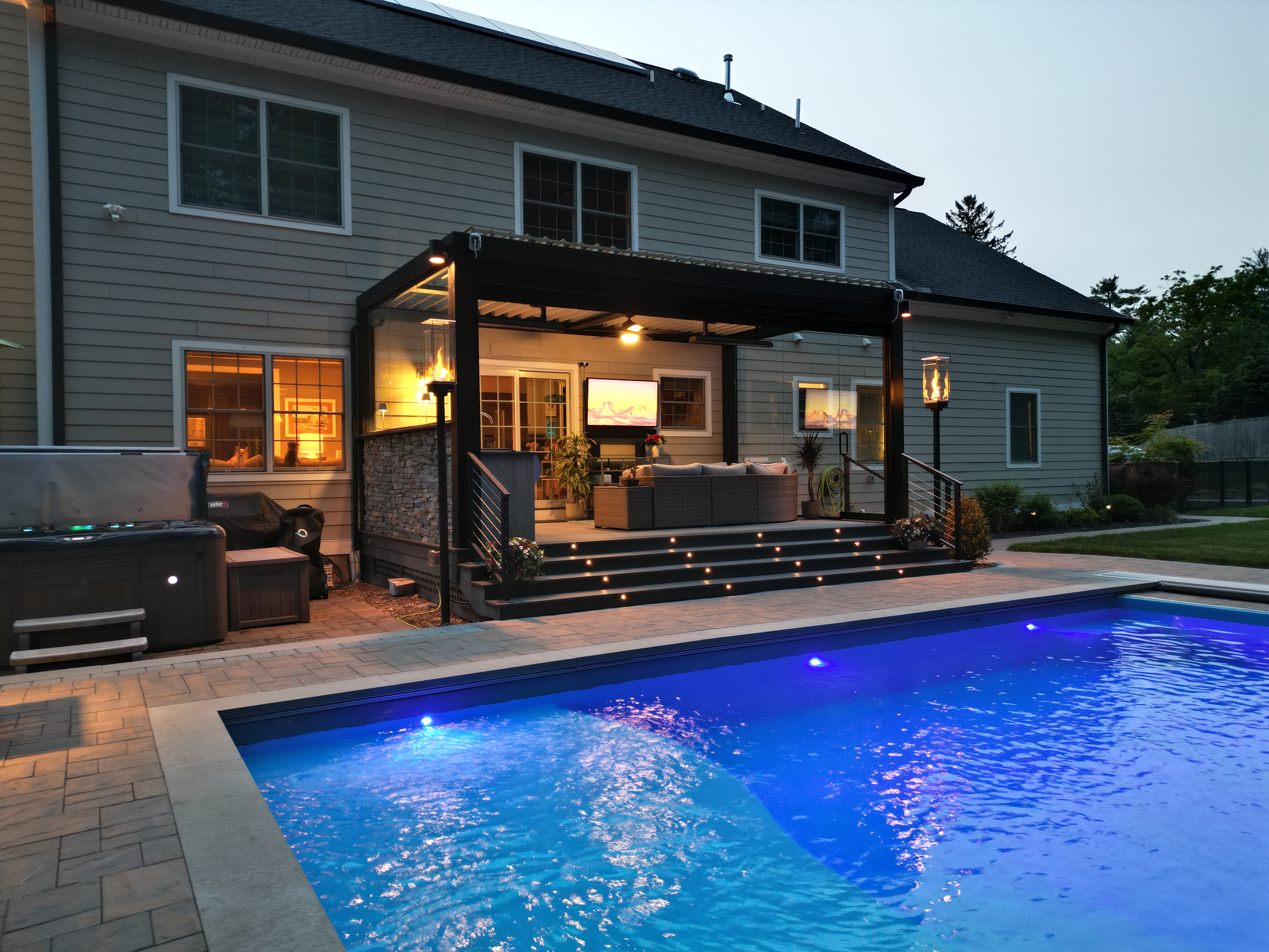 poolside deck with black louvered roof, glass side wall with door, outdoor kitchen, retractable screen, lounge seating, and step lighting