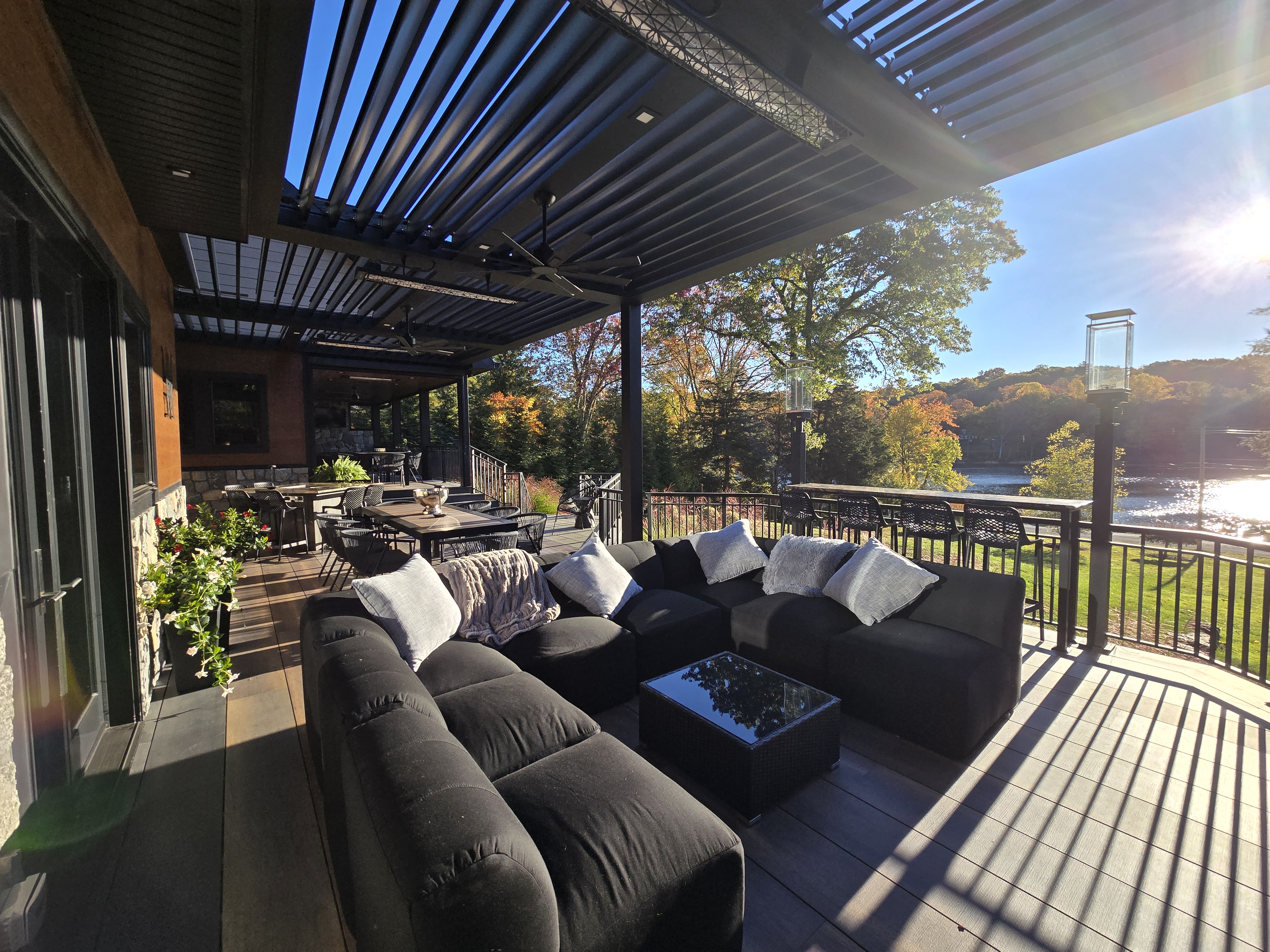 deck with louvered pergola, outdoor seating, and dining area overlooking a lake and colorful fall foliage