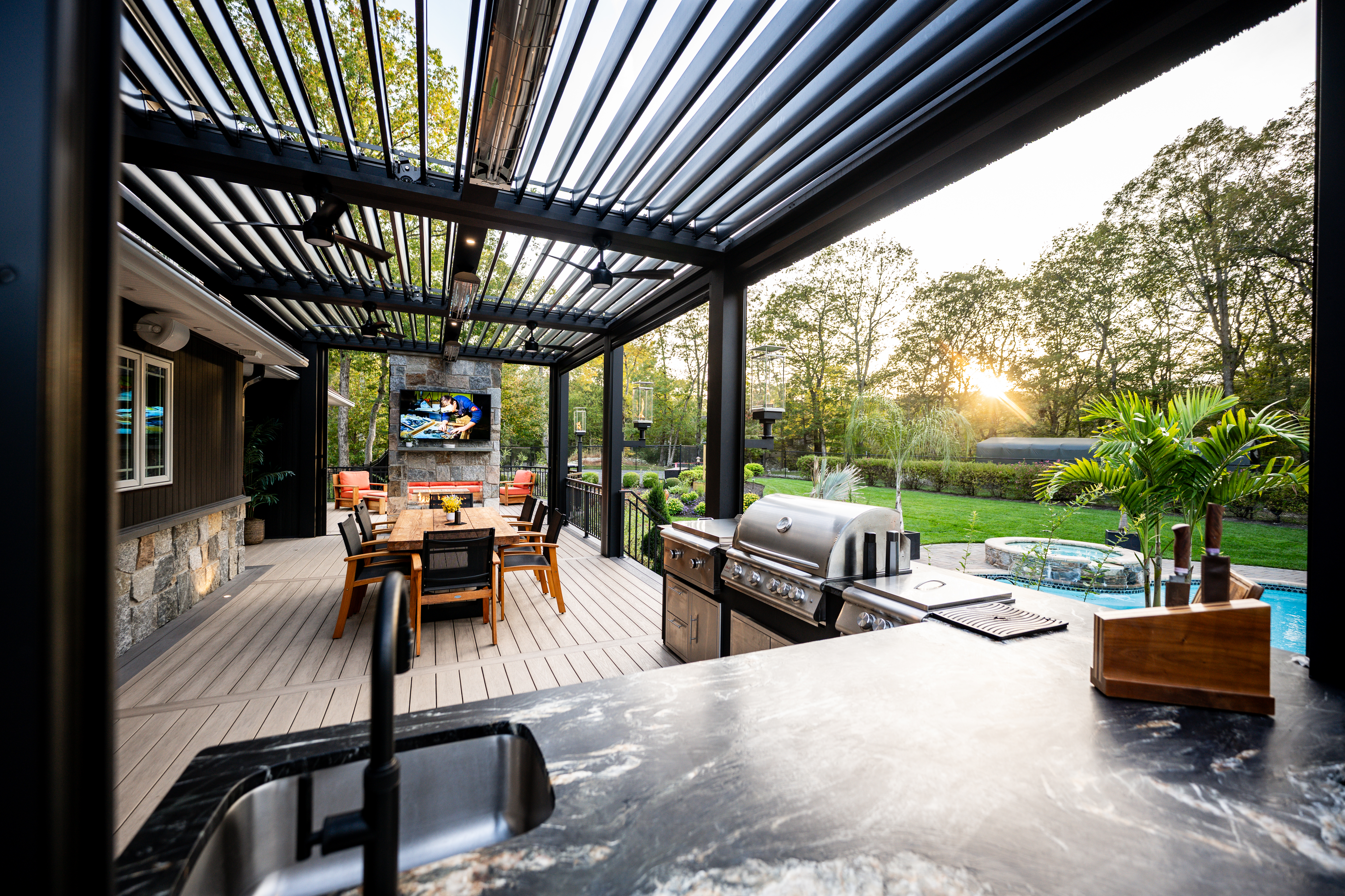 covered deck with outdoor kitchen, dining table, fireplace, and view of backyard pool and landscaping