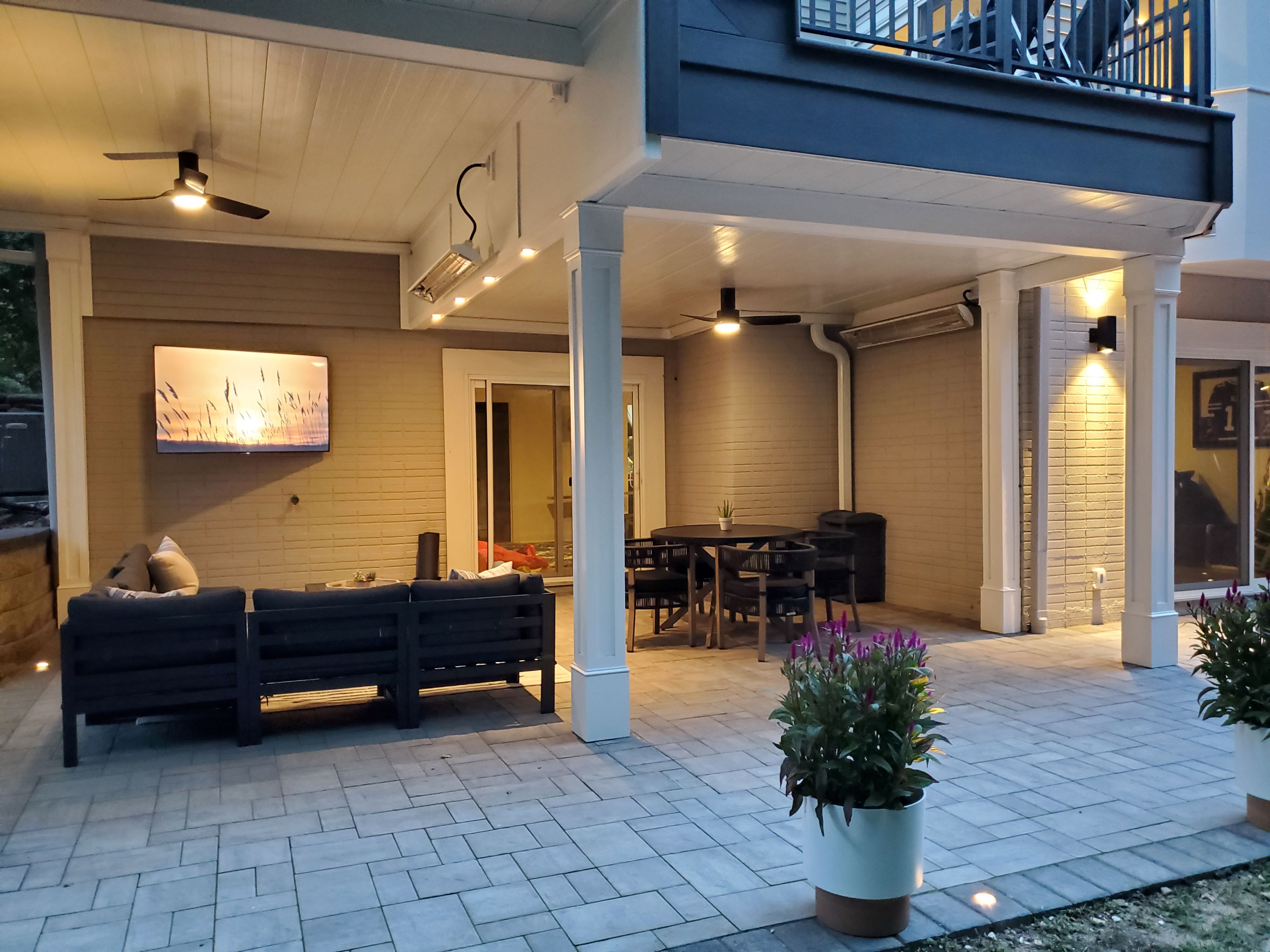 finished under deck space with two zones, lounge area with sectional sofa and wall-mounted TV on left, dining area with round table and chairs on right, white columns, ceiling fans, and paver flooring
