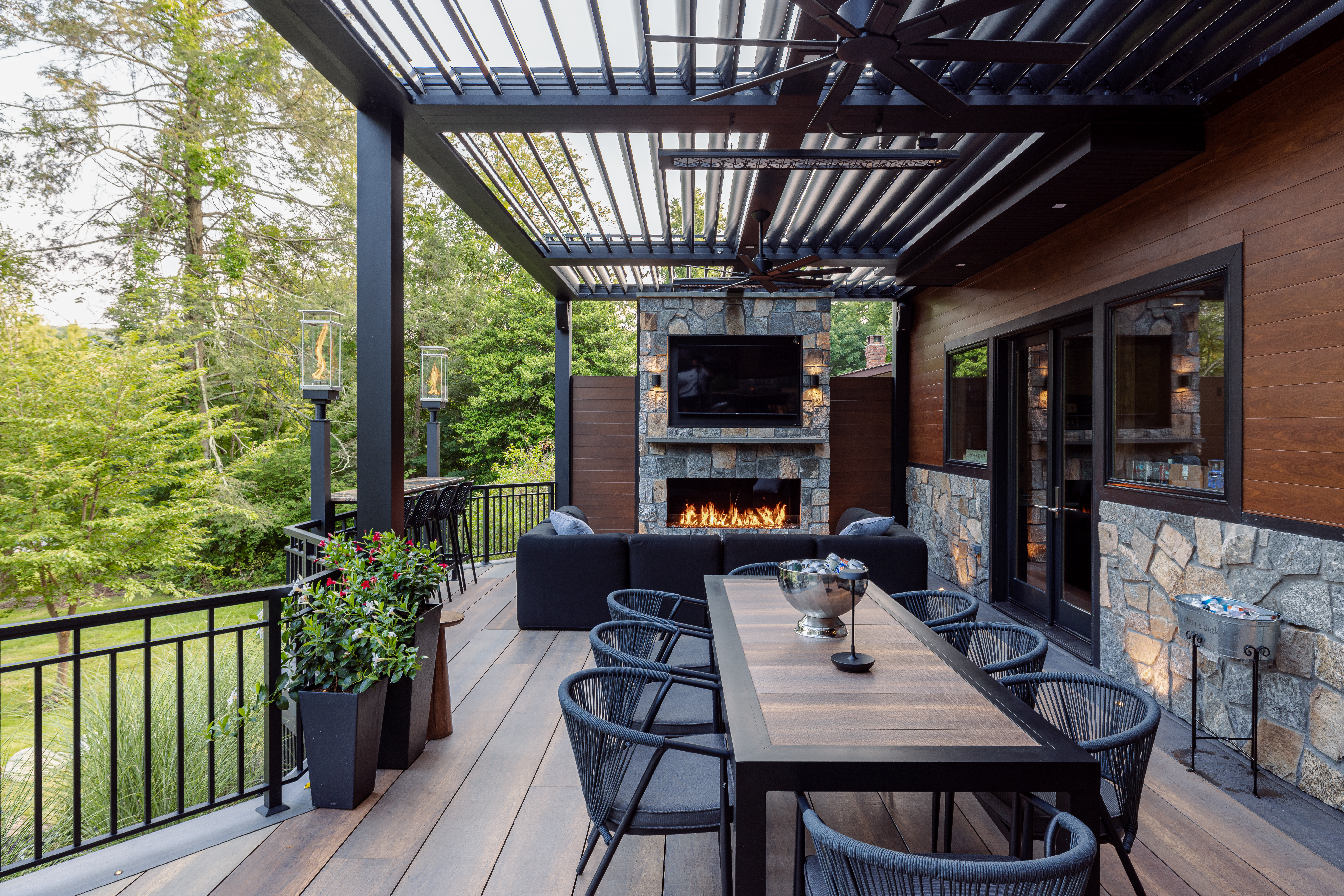 elevated deck with louvered roof, stone fireplace wall with outdoor TV, composite decking, ceiling fans, and rail-top bar seating overlooking backyard
