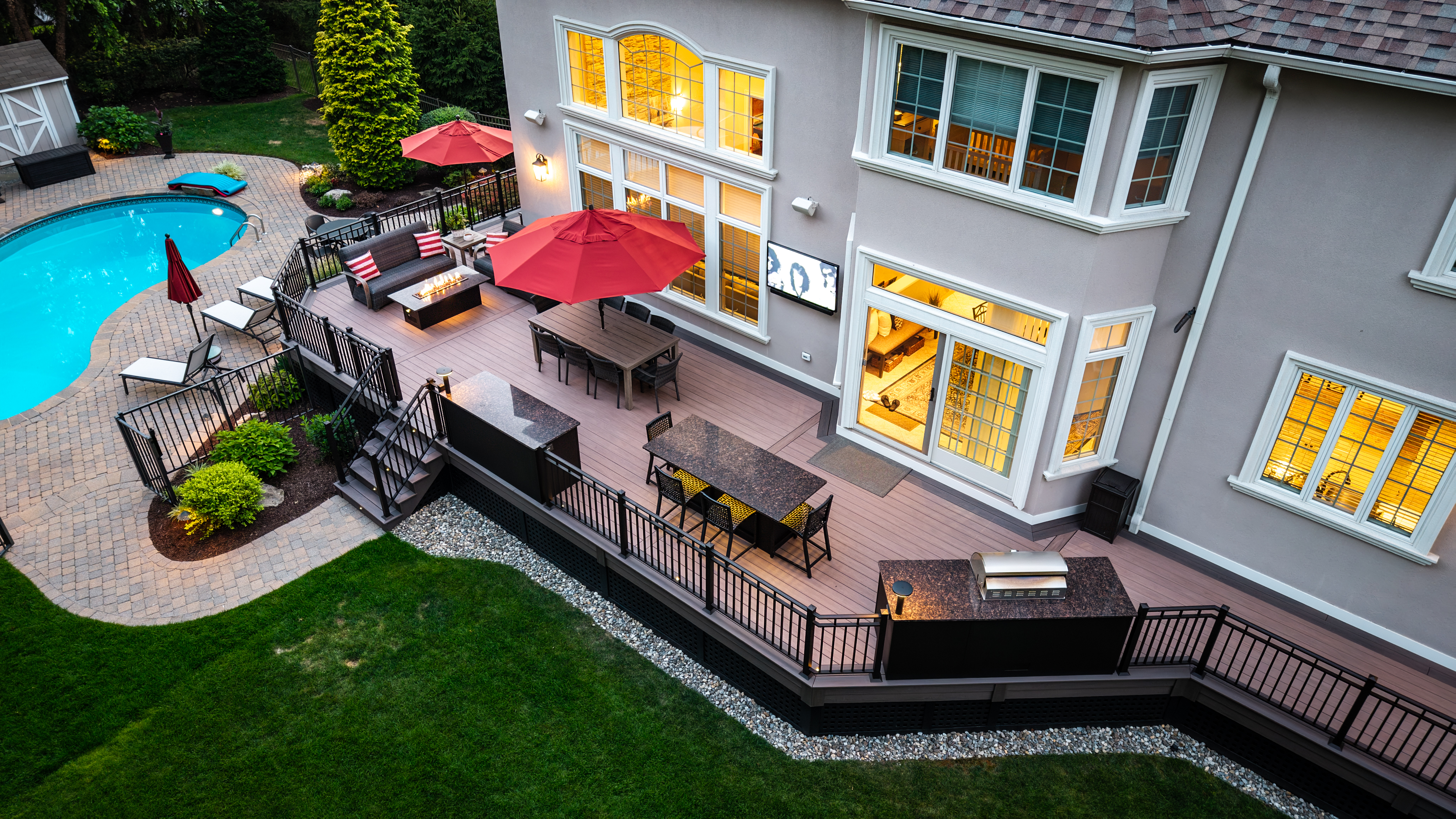 raised deck with outdoor dining tables, red umbrellas, built-in grill, and steps leading down to a poolside patio