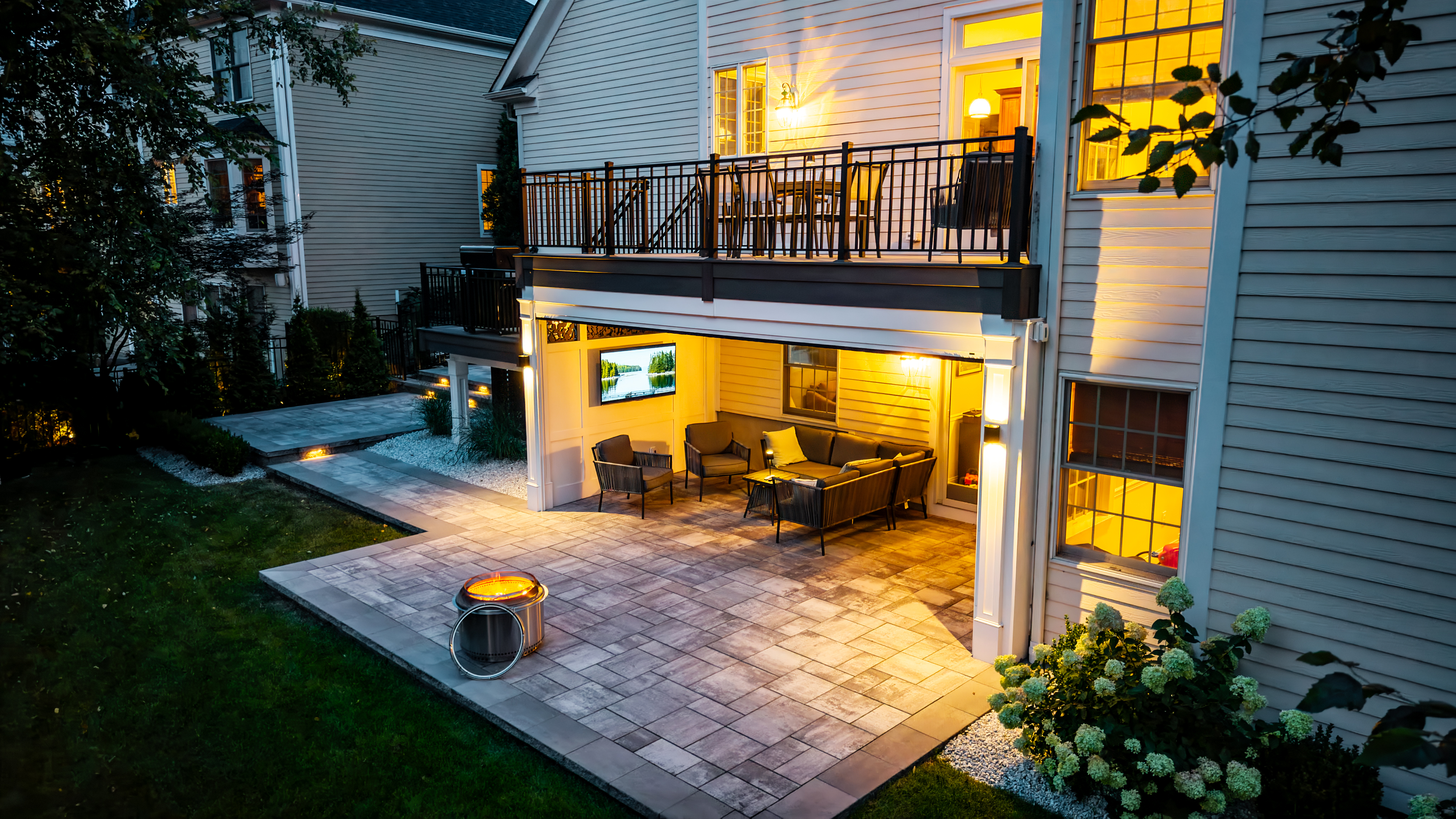 under-deck patio with built-in TV wall, lounge seating, modern fire pit, and paver hardscaping to maximize backyard space