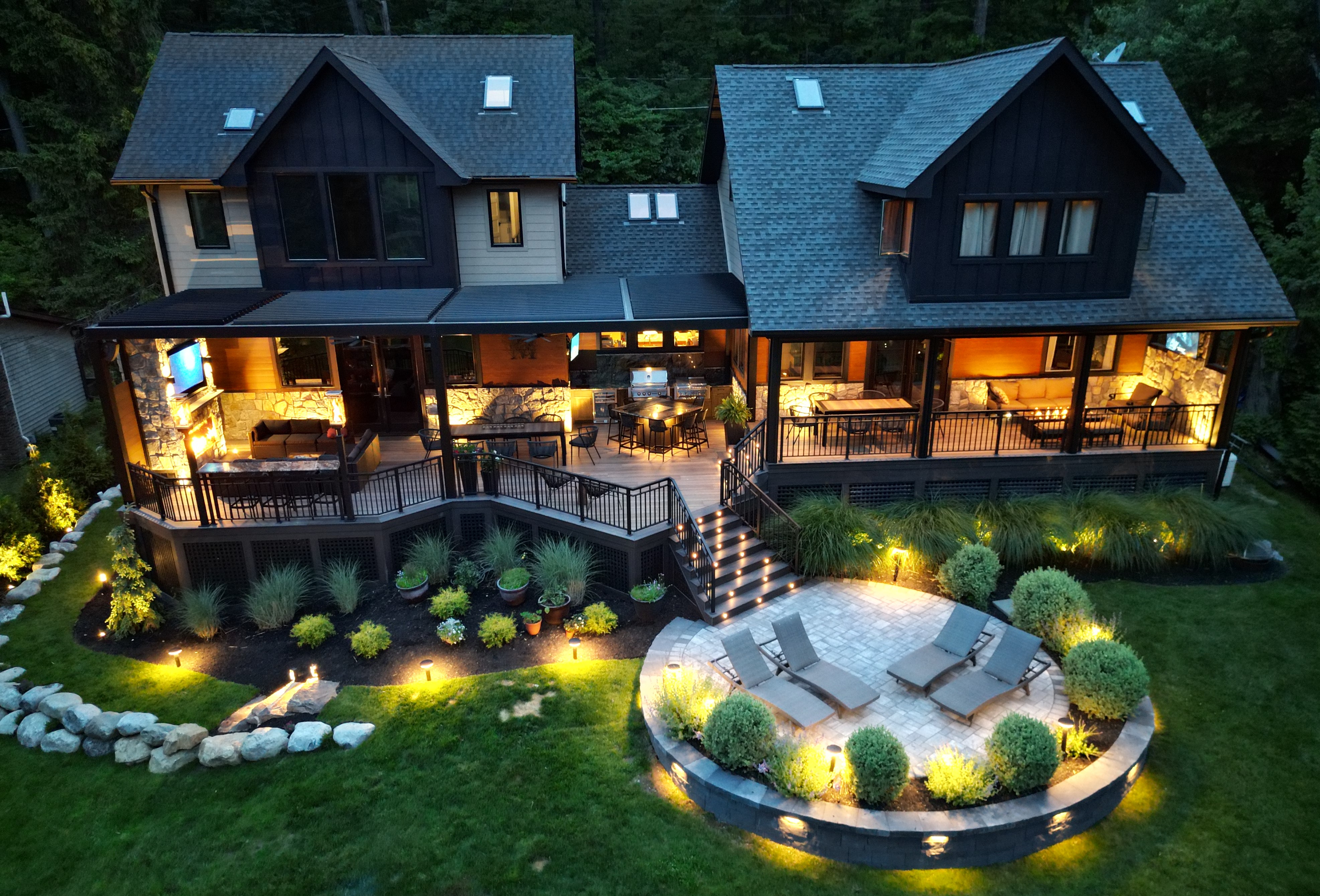 large covered deck with black louvered roof, outdoor kitchen, dining area, stone fireplace lounge, and lower patio with lounge chairs