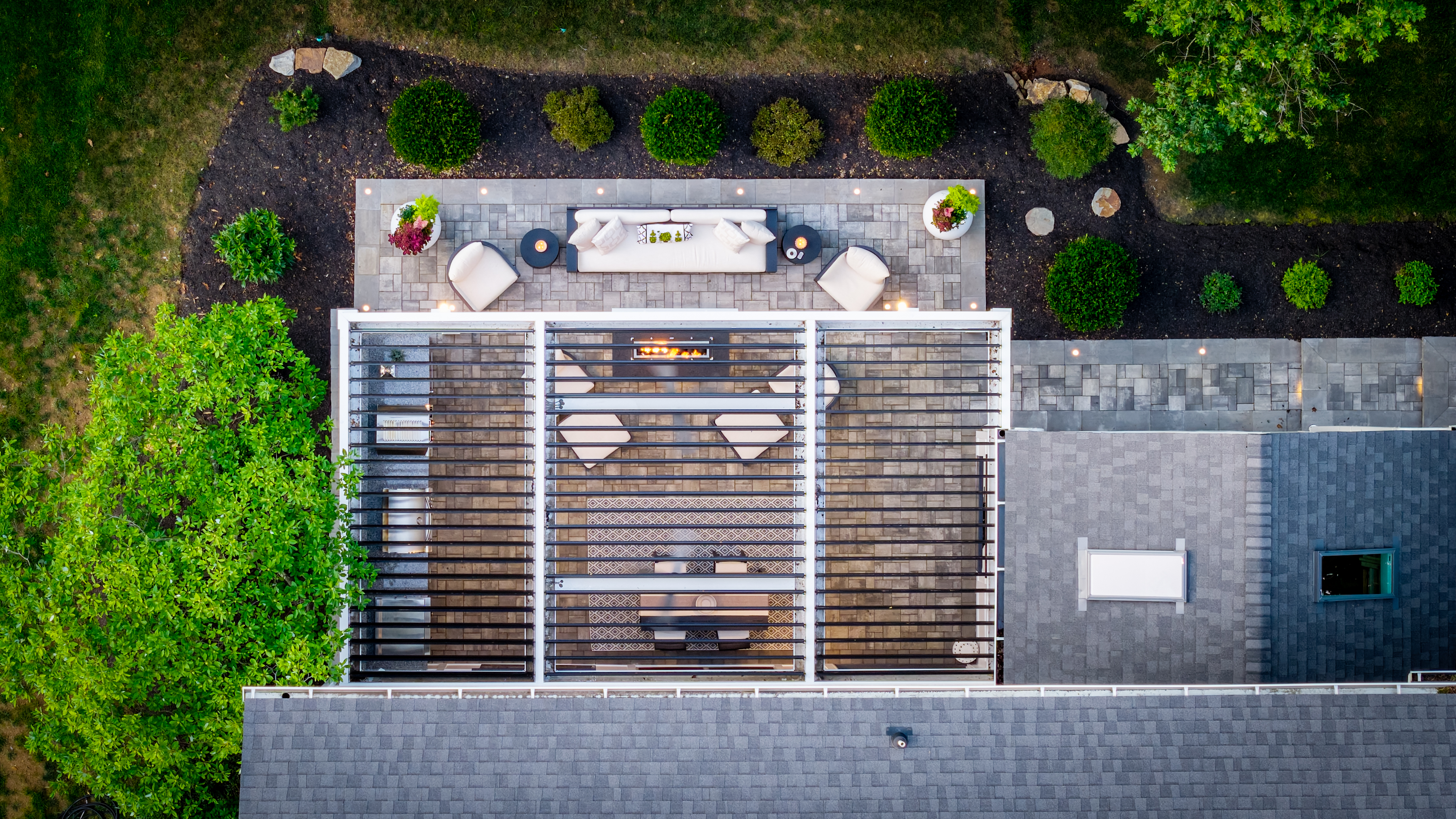 aerial view of compact backyard patio with louvered roof pergola, linear fire feature, outdoor lounge seating, and paver walkway with integrated lighting