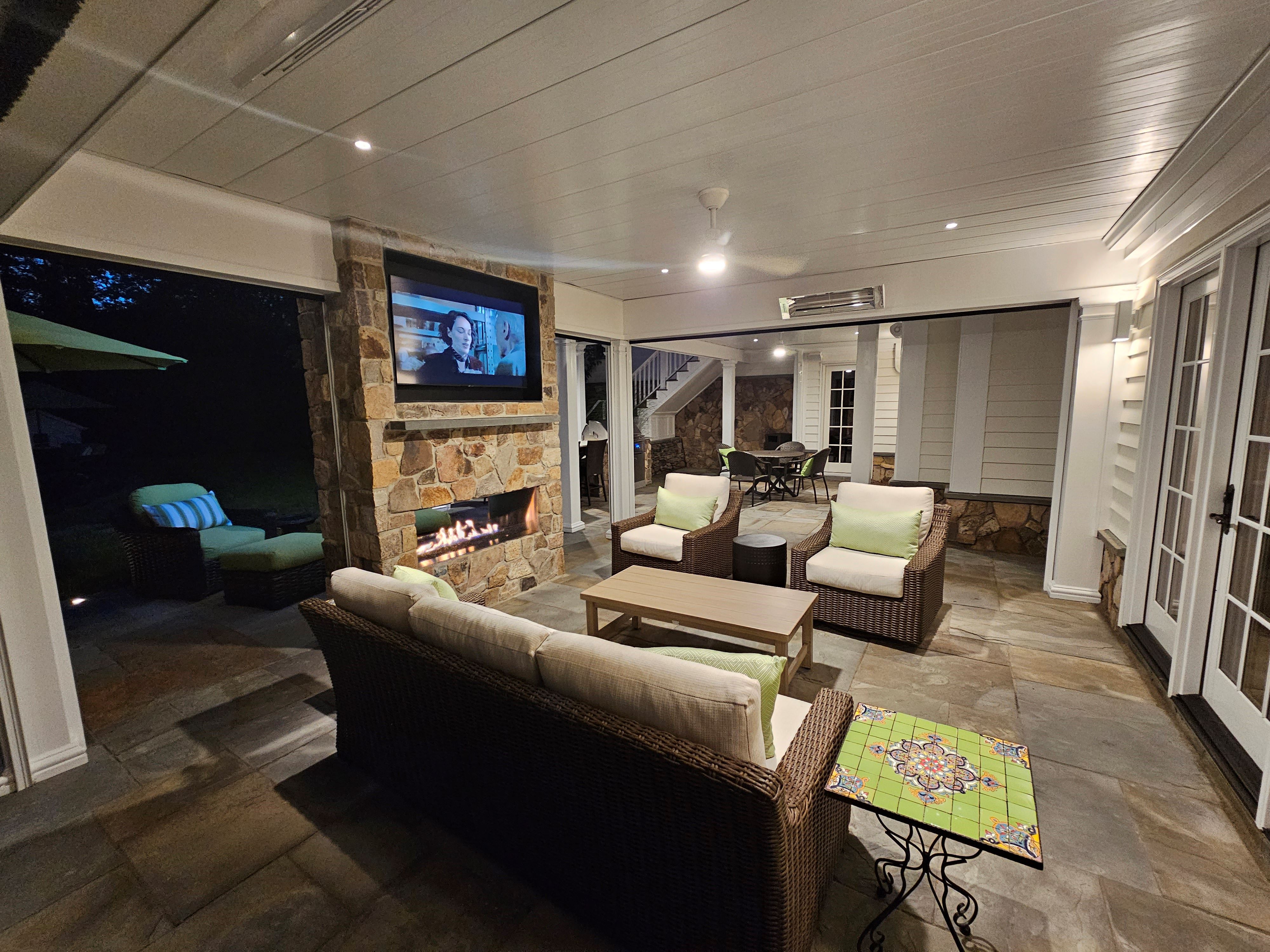 under deck lounge area at night with white paneled ceiling, stone fireplace, wall-mounted TV, wicker seating, infrared heater, flagstone floor, and French doors