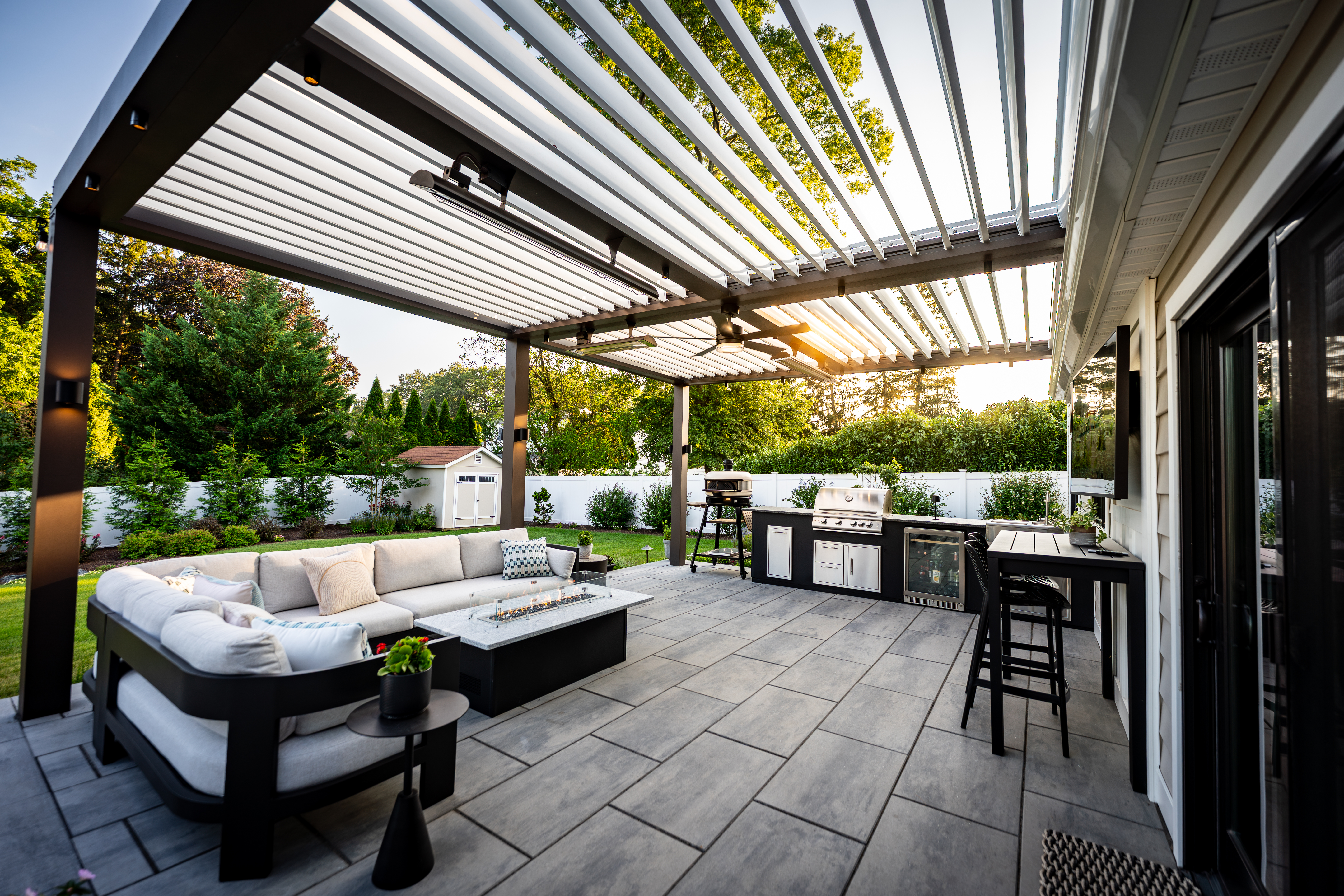 modern backyard patio with black motorized pergola, outdoor kitchen with built-in grill, sectional seating around fire table, and large paver flooring