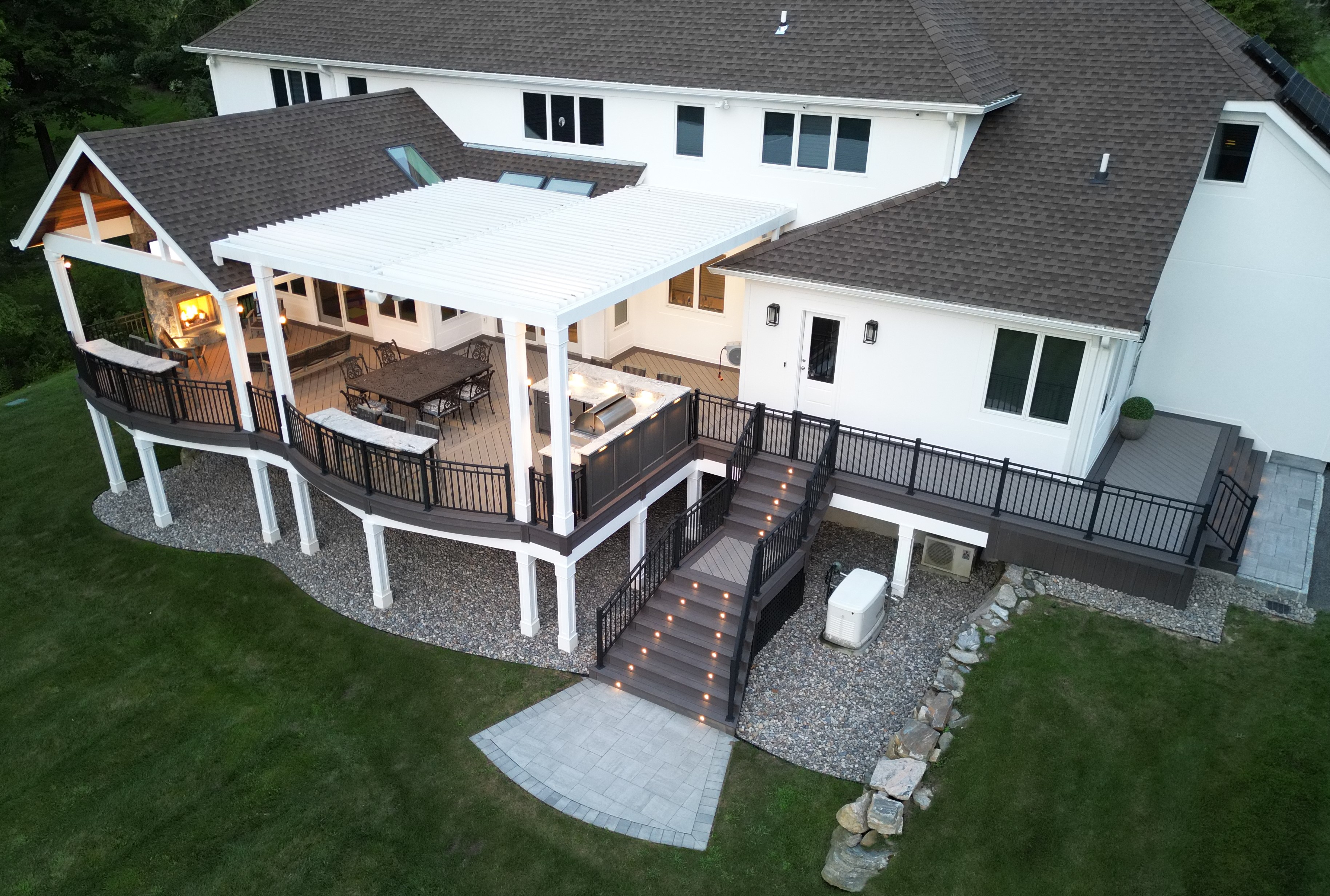 elevated deck with white louvered roof, outdoor kitchen, dining area, stone fireplace lounge, and step lighting