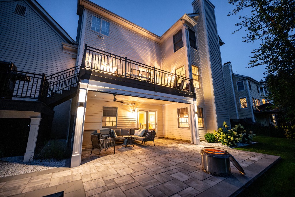 Raised deck with black railing above covered patio featuring outdoor seating, ceiling fan, and stone paver flooring illuminated at dusk, Made by Deck Remodelers