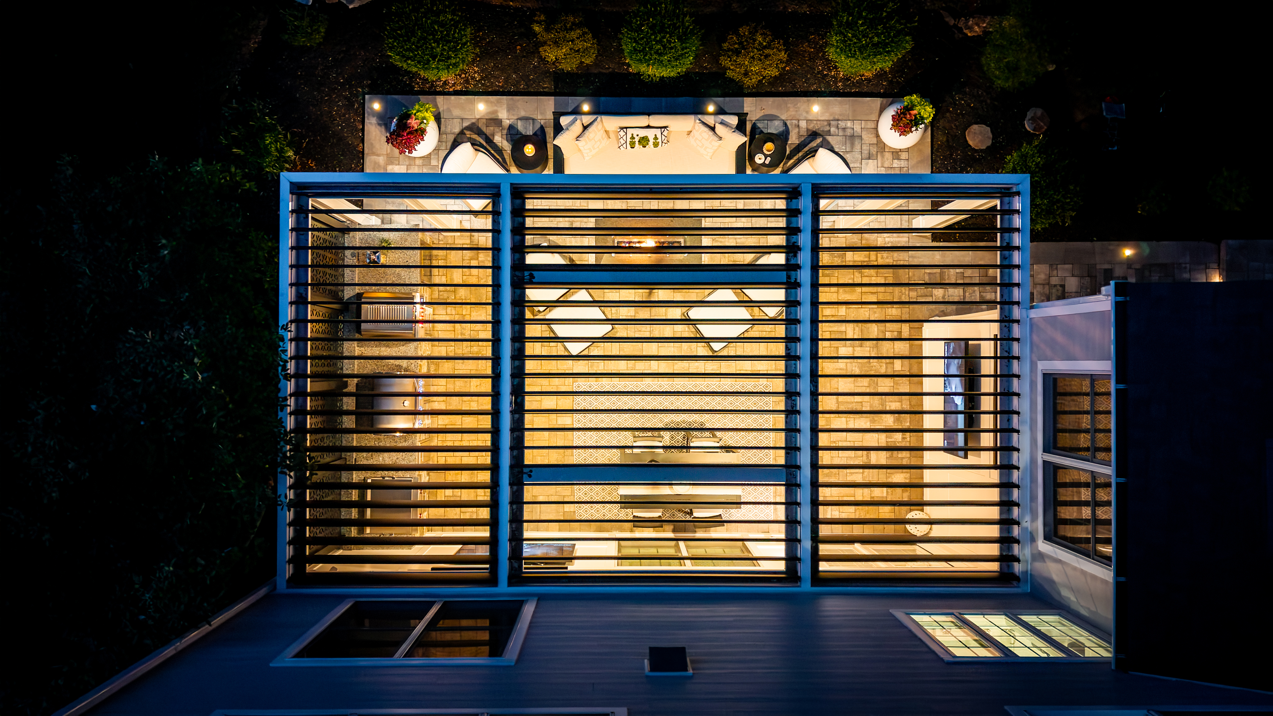 aerial view of modern backyard patio with black louvered roof, outdoor kitchen, lounge seating, and illuminated paver flooring at night