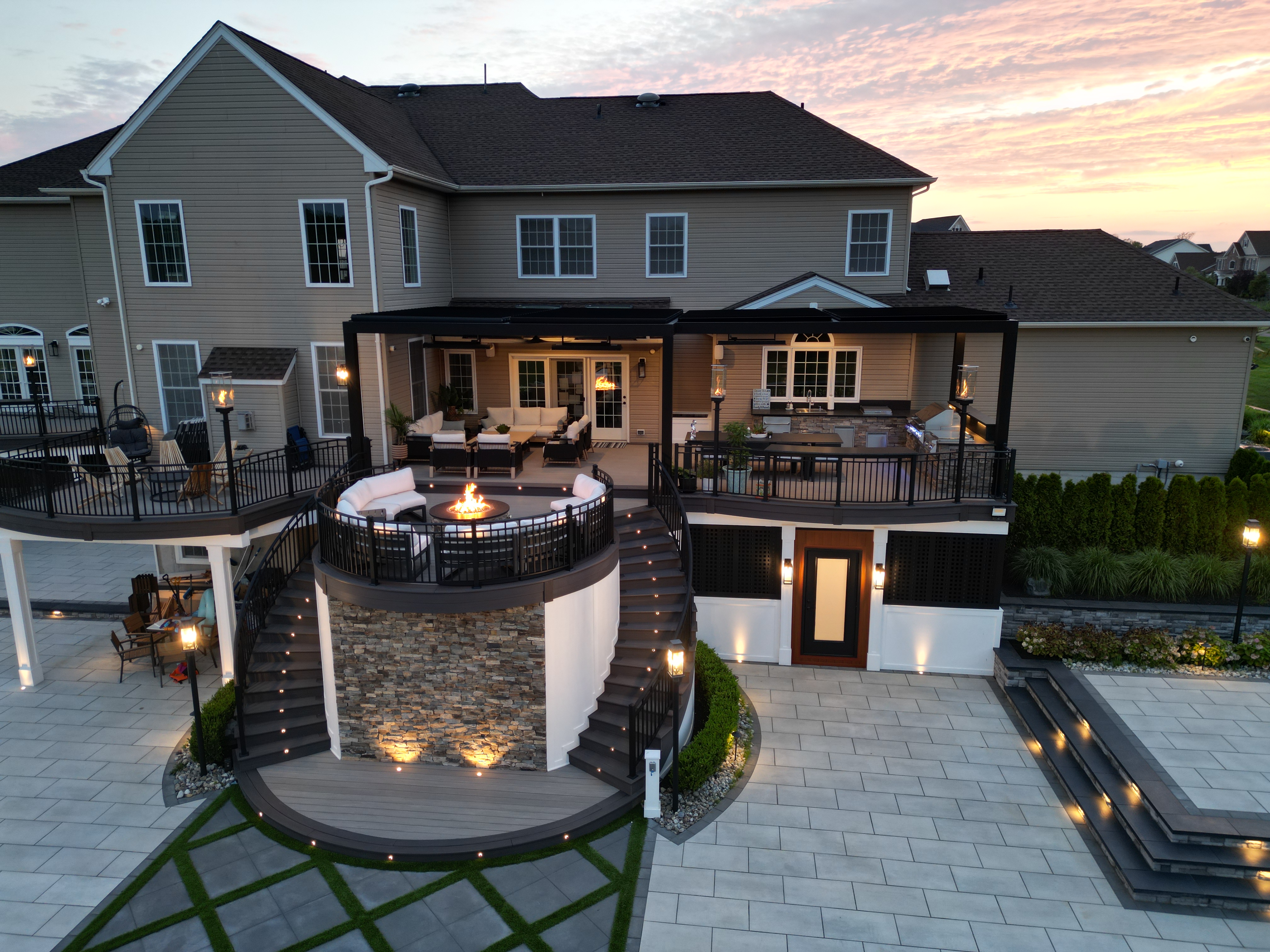 multi-level backyard deck with louvered roof, curved fire pit lounge, outdoor kitchen, stone stair risers, and expansive paver patio with step lighting
