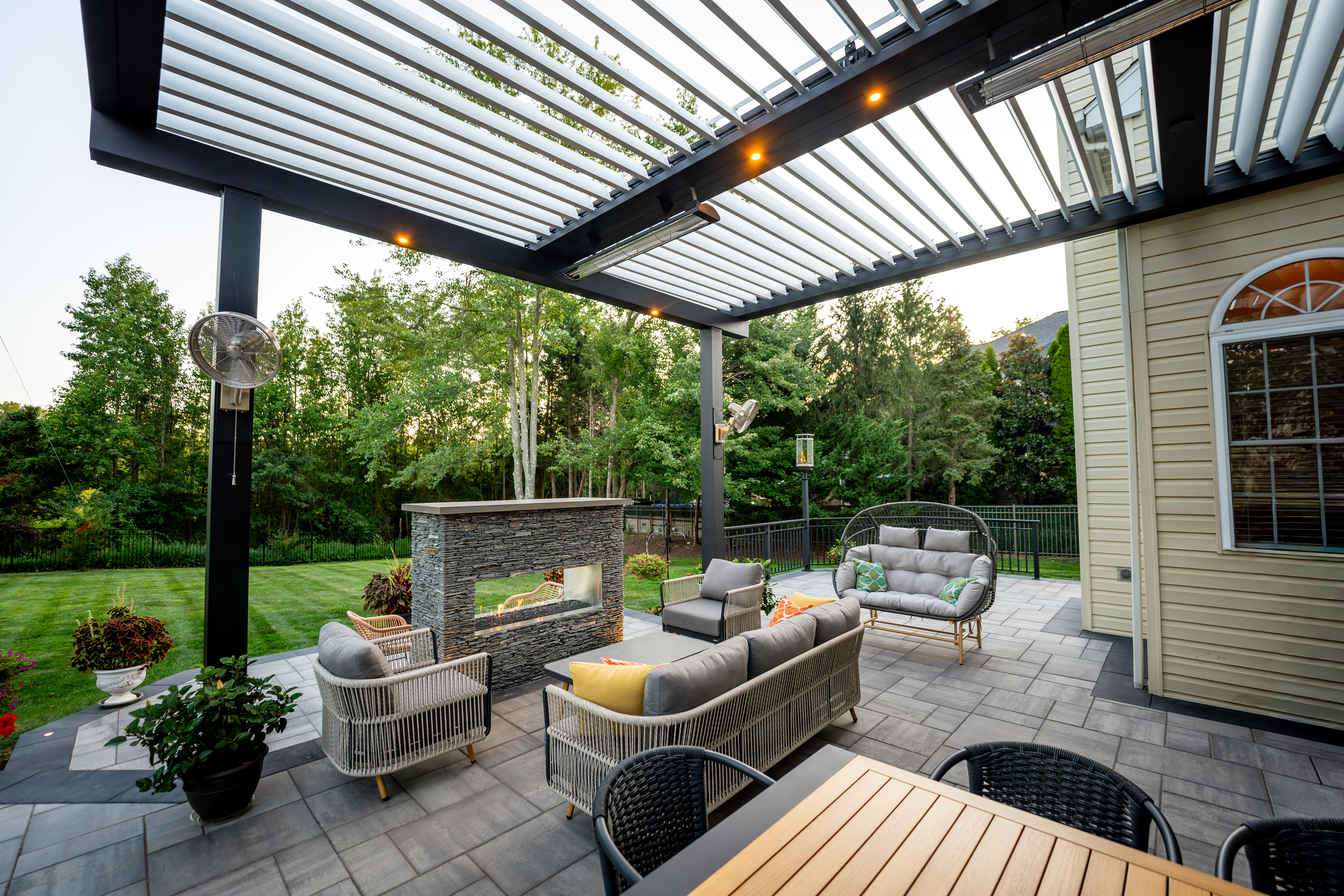 patio with louvered pergola, stone fireplace, and outdoor seating surrounded by trees and greenery