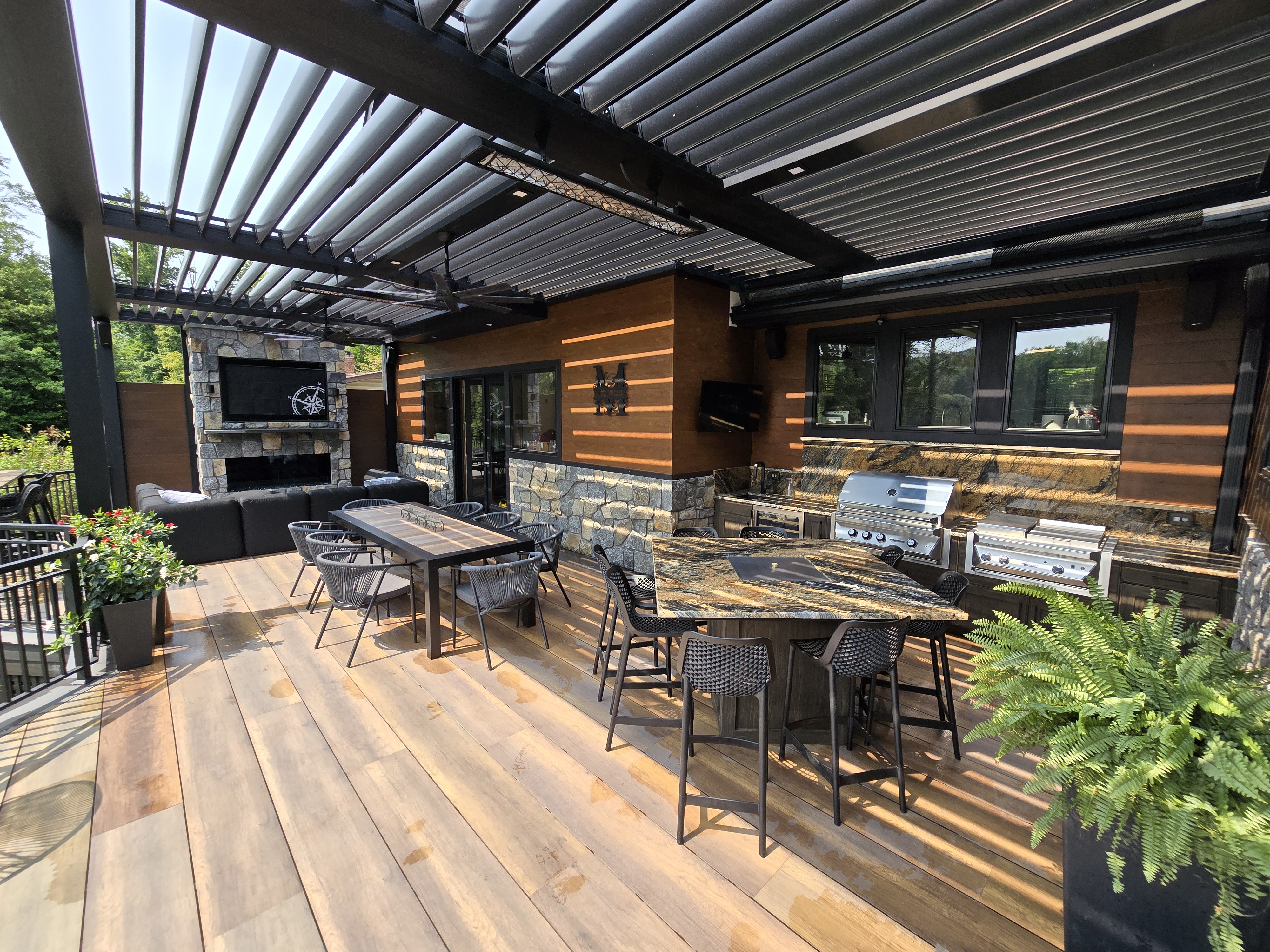 sunlight streaming through black louvered roof onto deck with stone fireplace lounge, granite outdoor kitchen, bar seating, and dining table