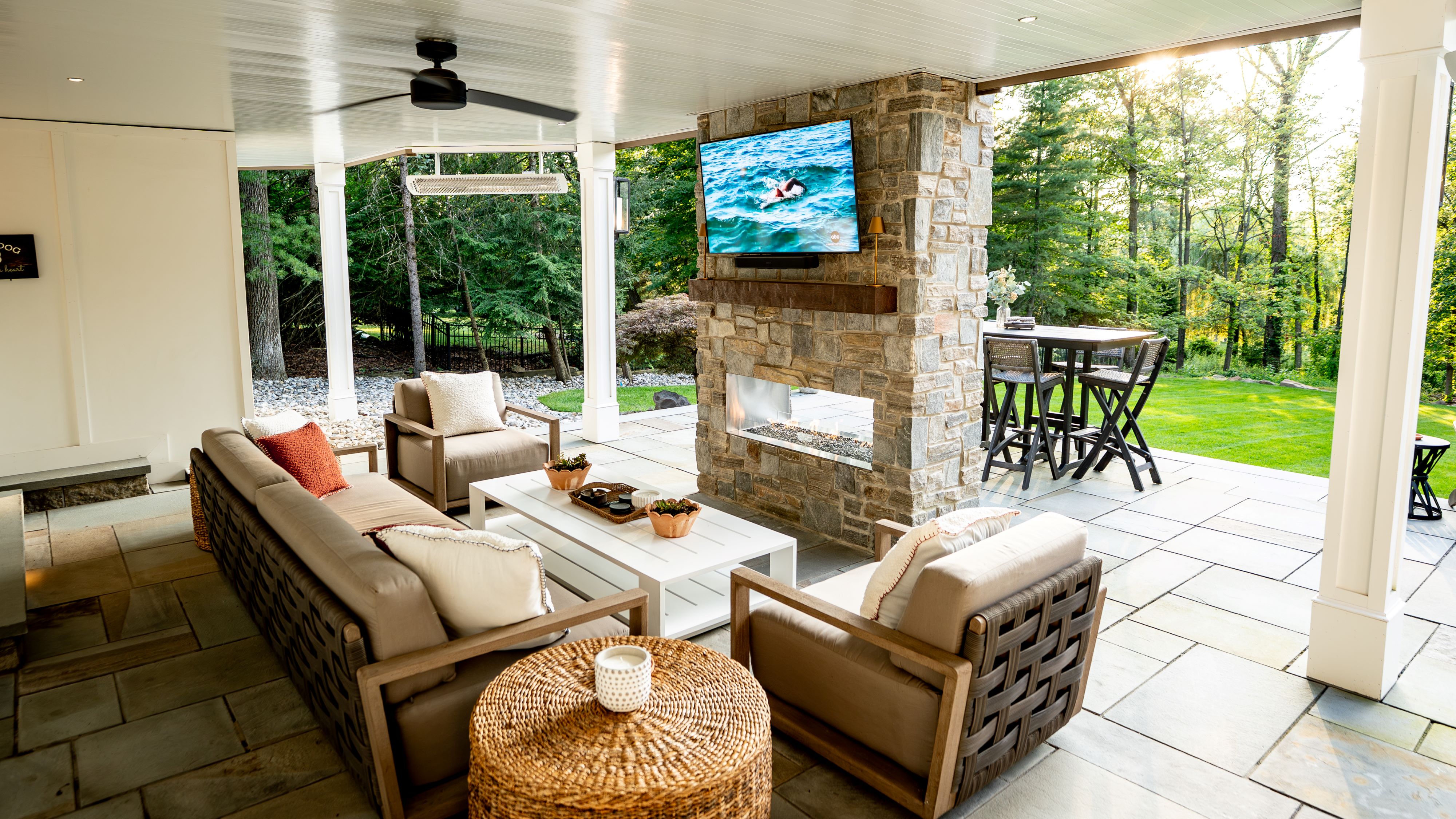 open under deck lounge with white paneled ceiling, stone fireplace, wall-mounted TV, lounge seating, wicker coffee table, flagstone flooring, and white columns opening to wooded backyard