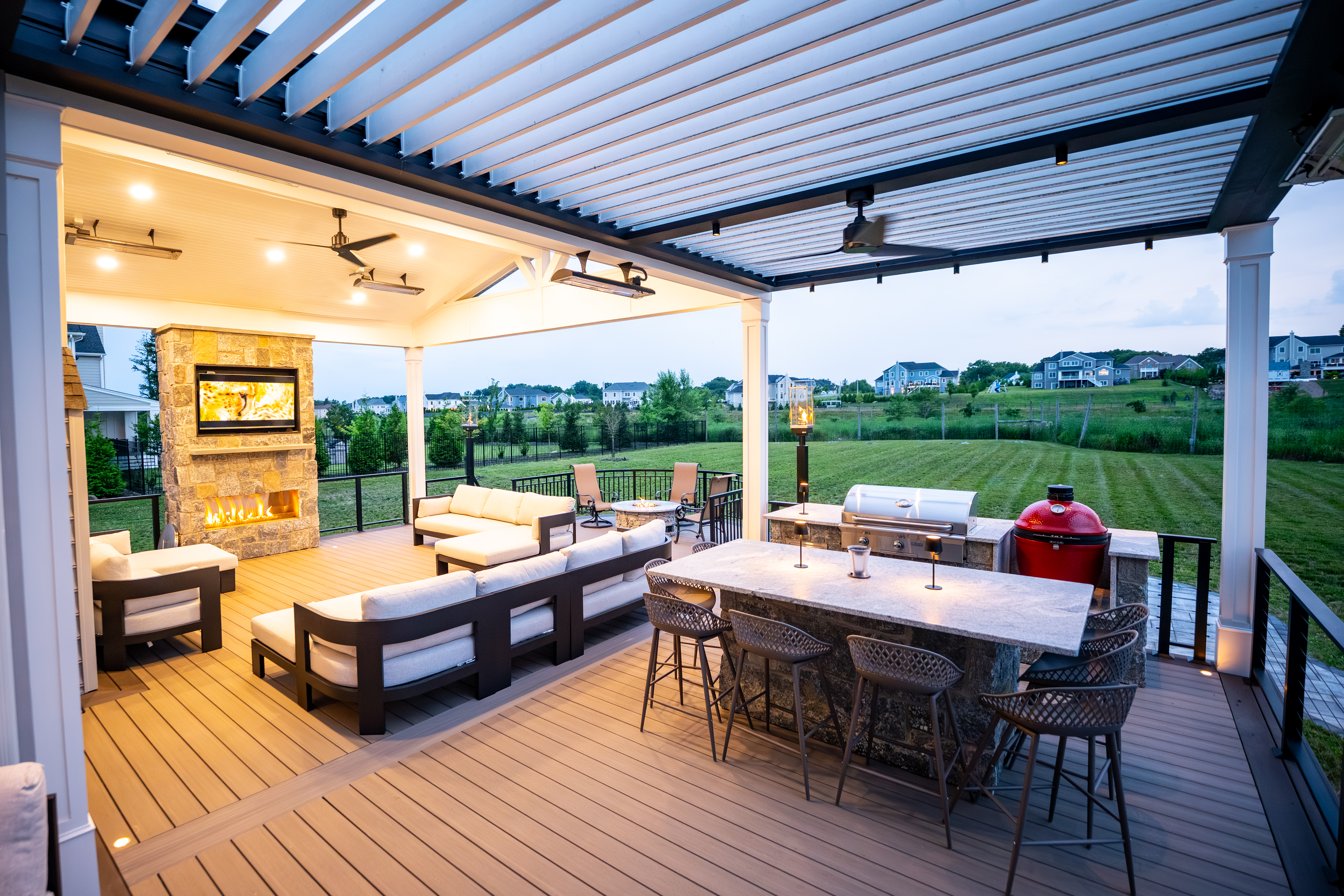 covered deck with louvered pergola, outdoor kitchen, bar seating, and stone fireplace with TV above