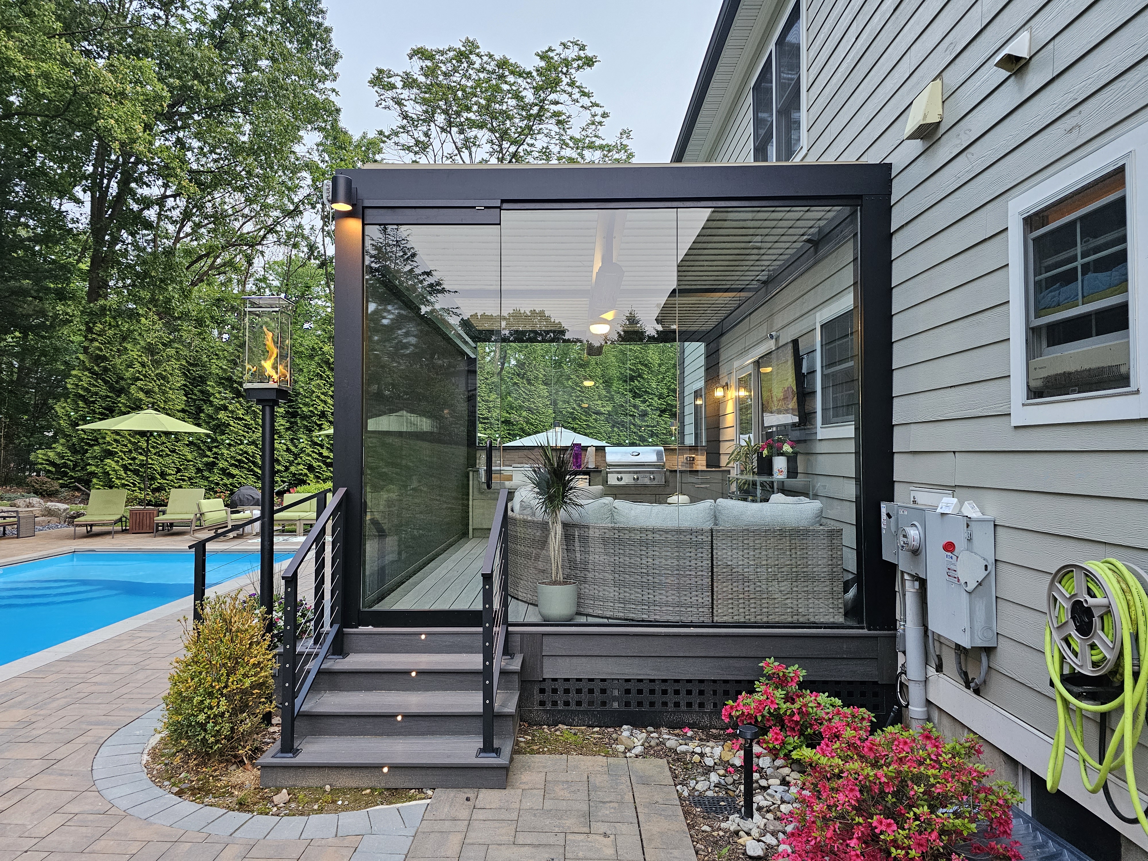 modern enclosed deck with glass walls under black louvered roof, outdoor sofa seating, and poolside view