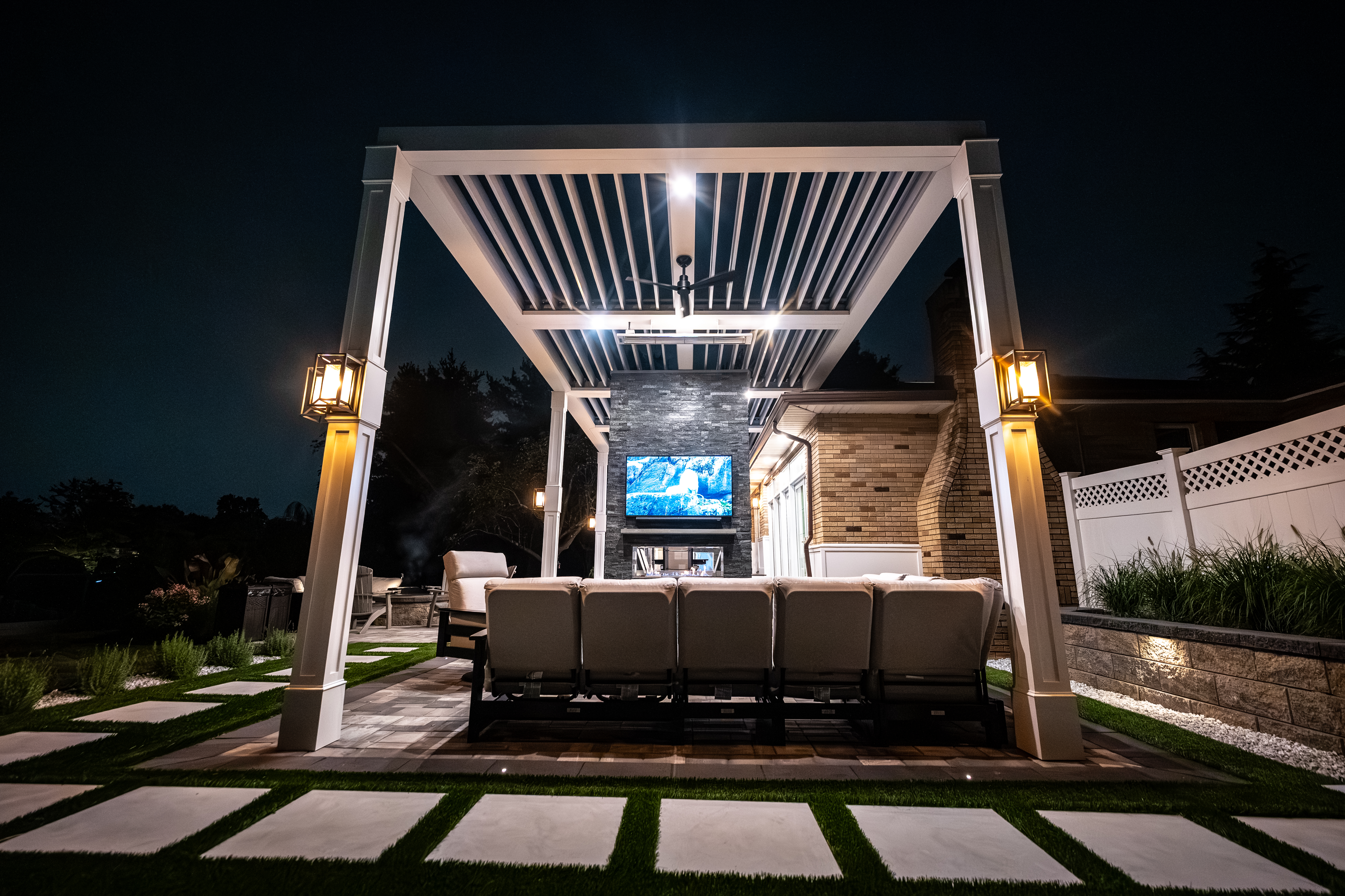 nighttime backyard with white louvered roof, stone fireplace wall with mounted TV, cushioned seating, and modern lantern lighting