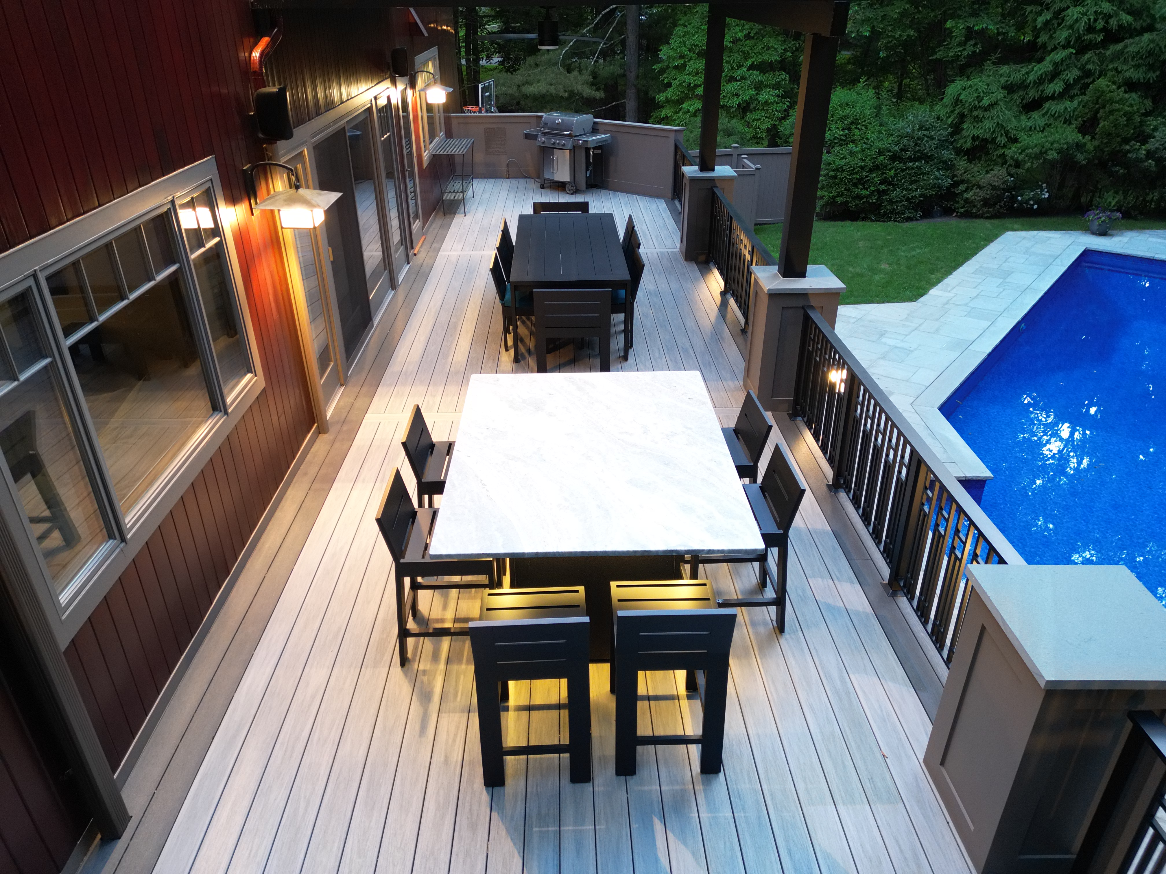 PVC poolside deck with marble-top dining tables, custom railings, accent lighting, and built-in grill station along a modern home exterior