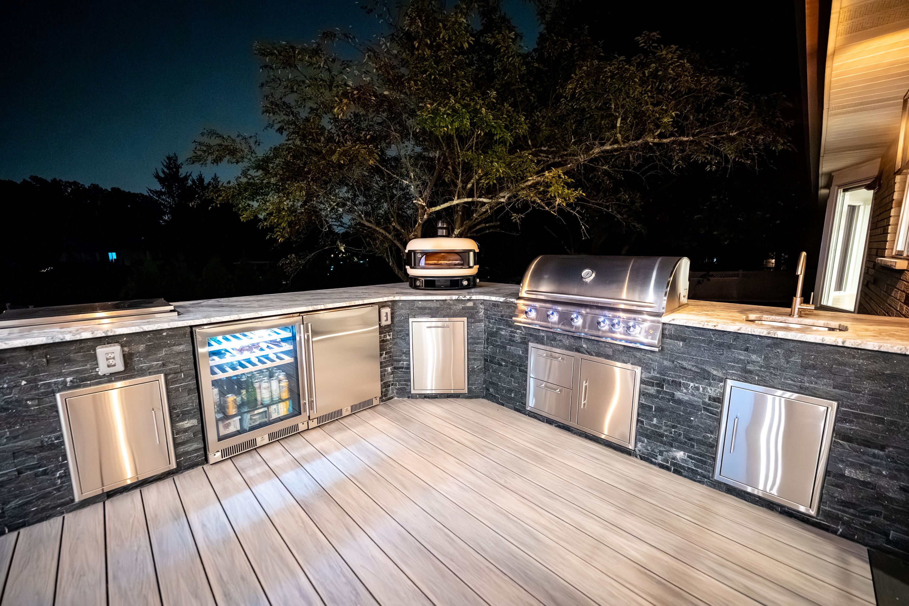 nighttime outdoor kitchen with built-in grill, pizza oven, beverage refrigerator, stone veneer cabinetry, and marble countertops on a deck