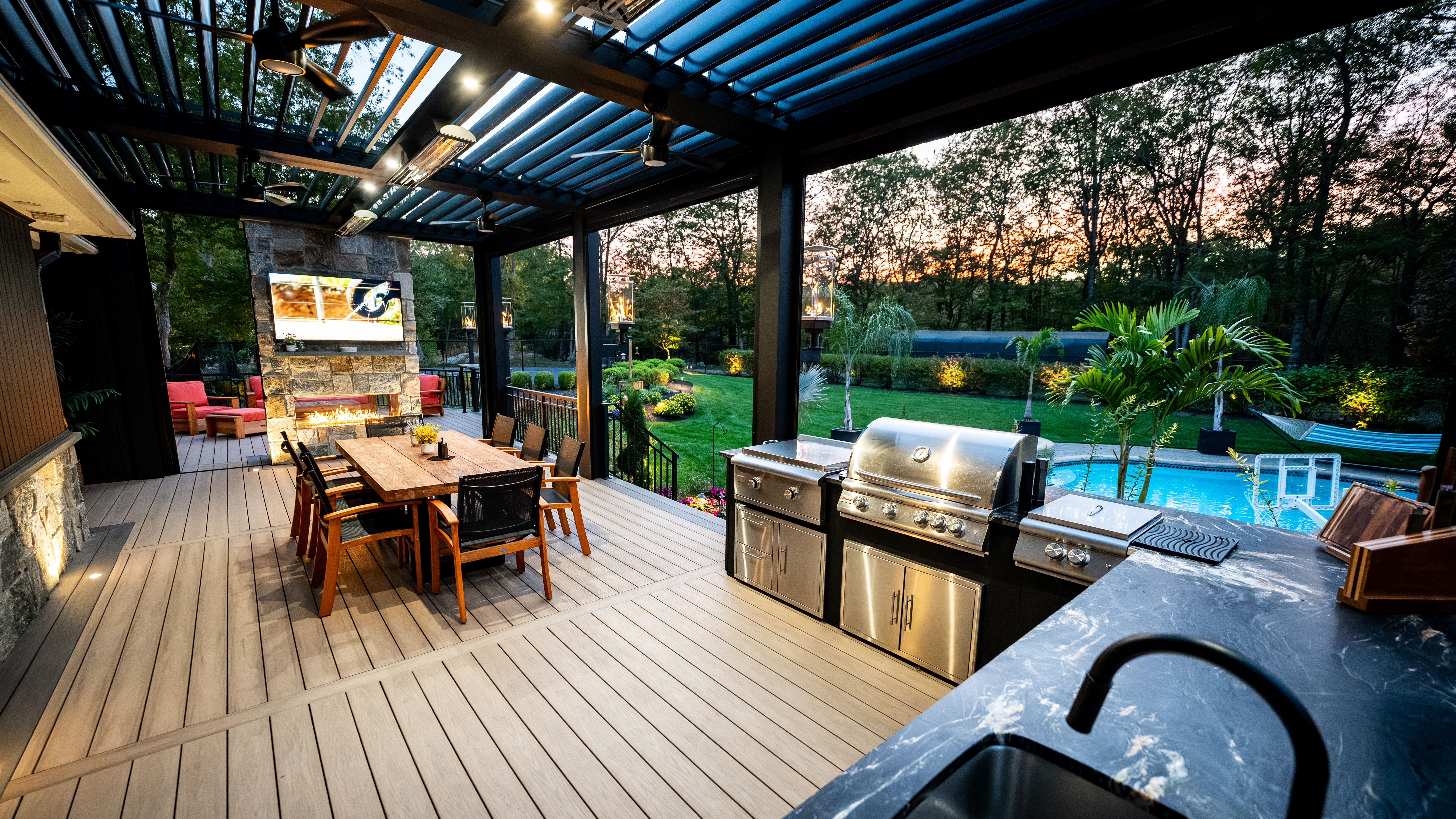covered deck with outdoor kitchen, dining area, and fireplace overlooking a backyard pool at sunset
