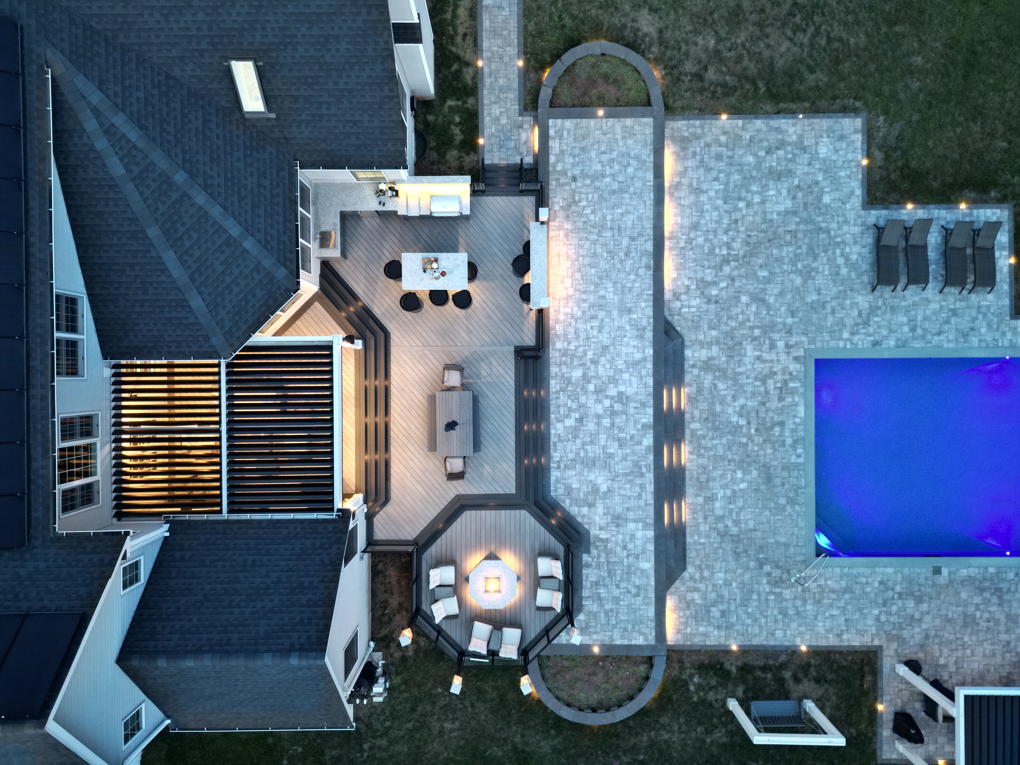 aerial view of backyard with black louvered roof on spacious deck, fire pit lounge, outdoor kitchen island, and illuminated pool