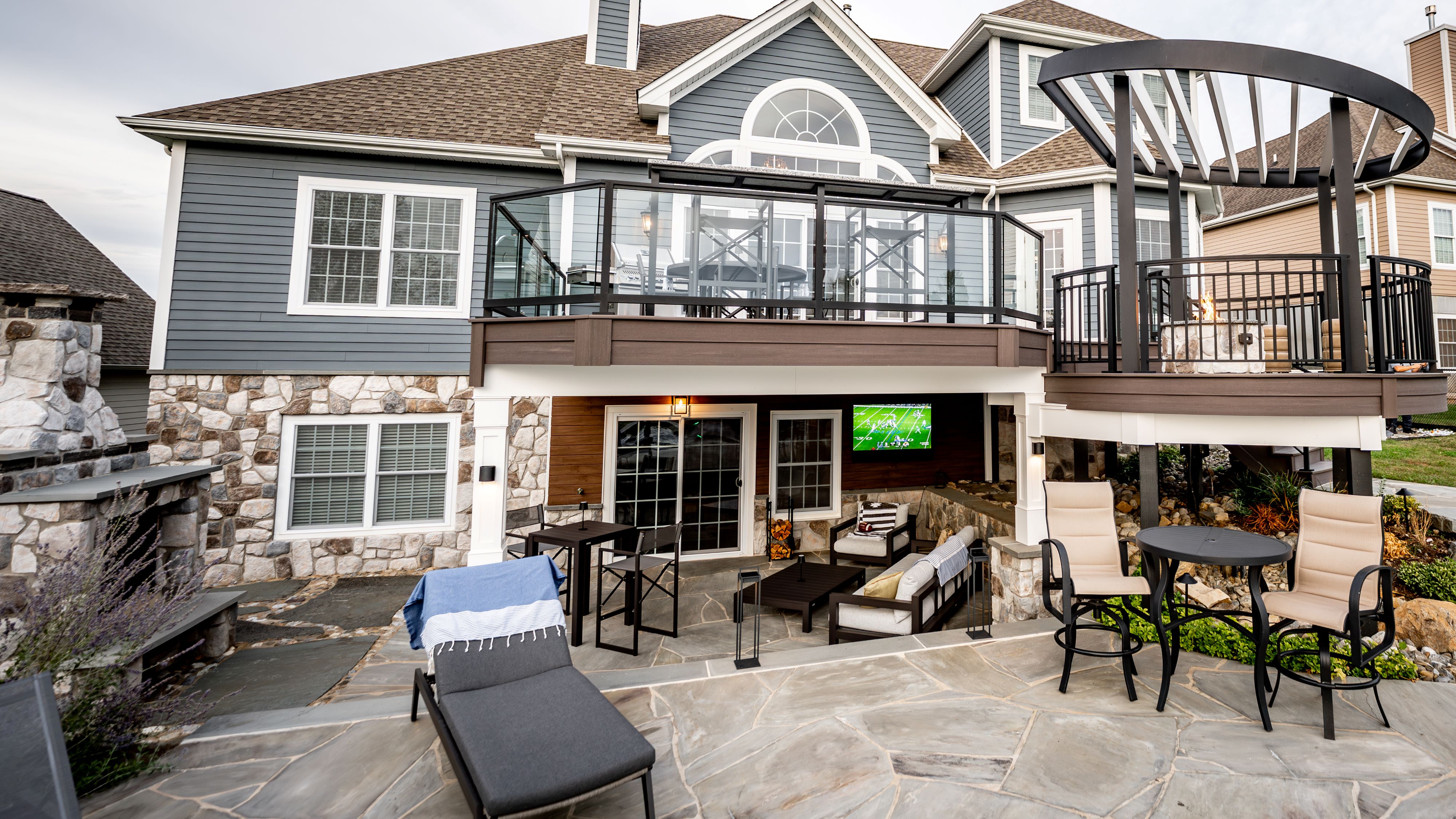 finished under deck lounge with wood ceiling, stone accent wall, wall-mounted TV, and lounge seating opening to a flagstone patio with bar-height table and chaise lounge