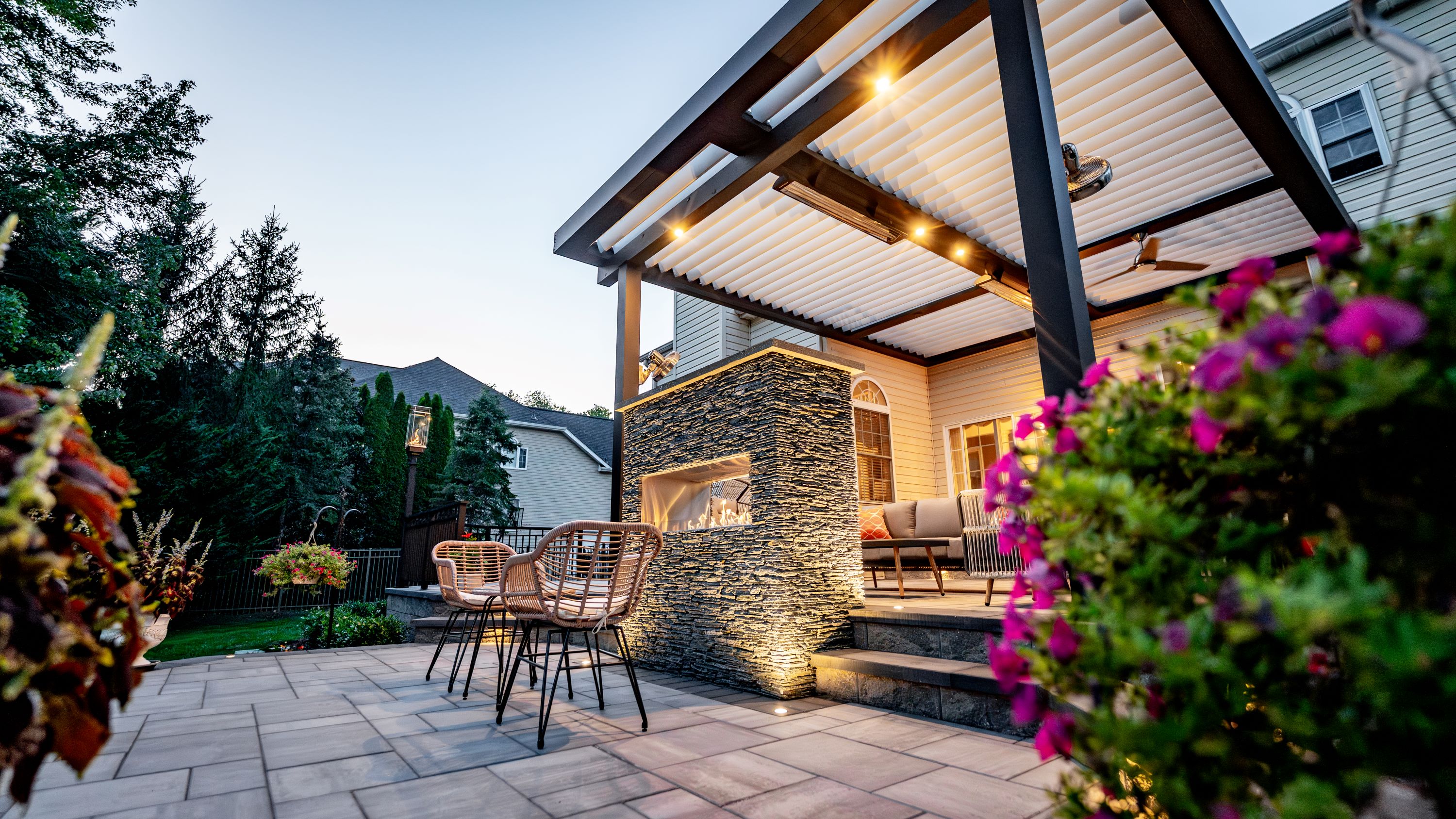 raised paver patio with louvered roof, double-sided stacked stone fireplace, lounge seating, and lighted steps