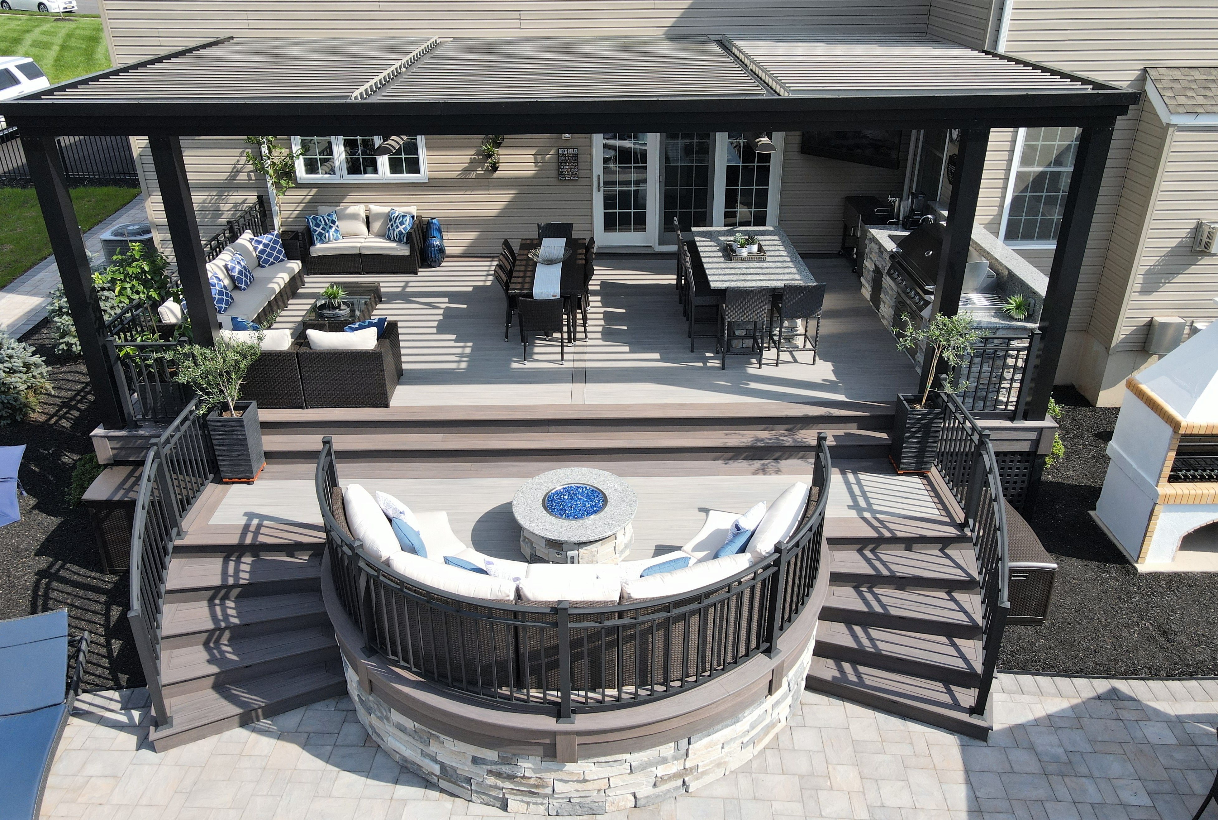 multi-level deck with louvered roof pergola, outdoor kitchen, dining area, curved fire pit lounge, and wide stairs cascading into a paver patio