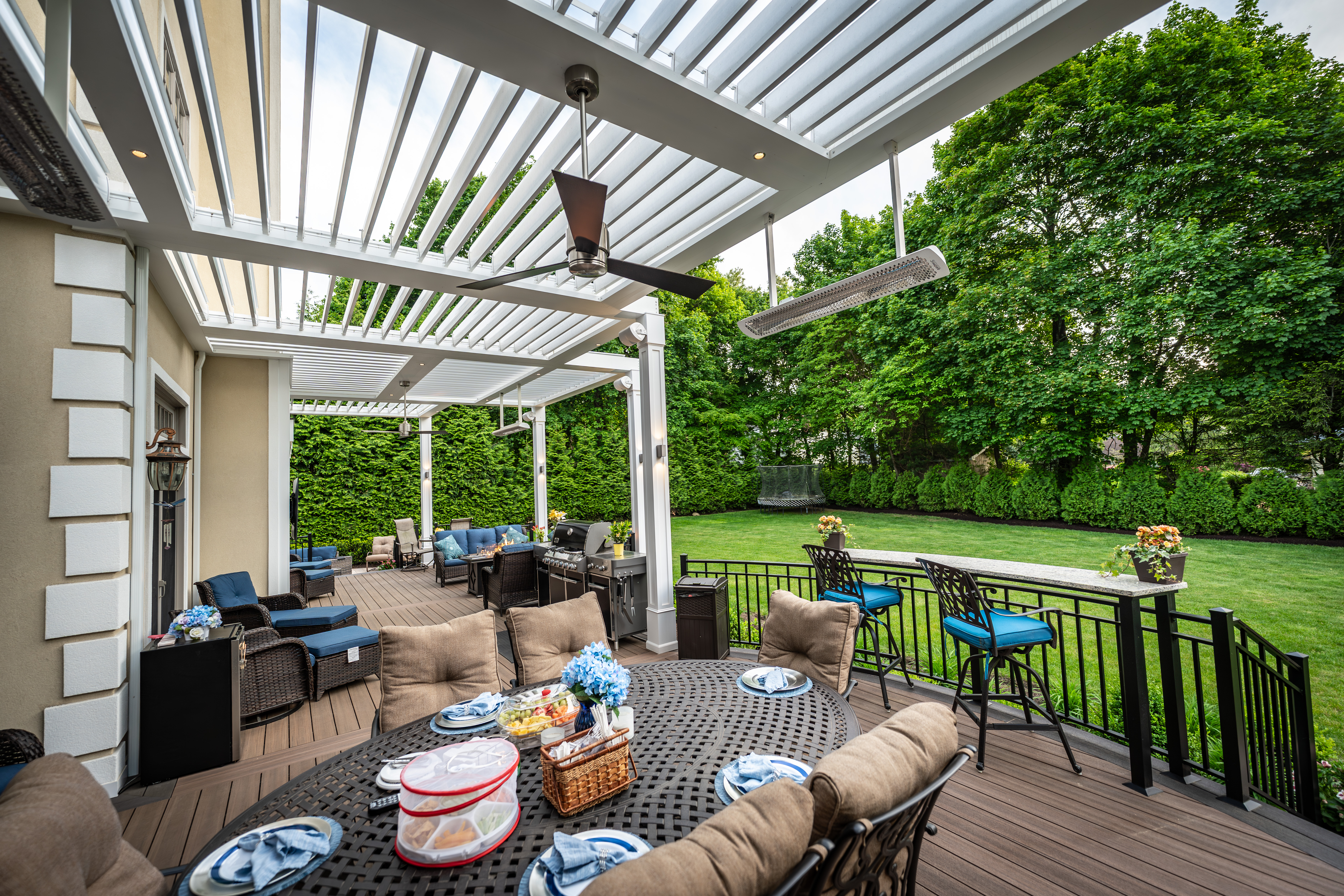 backyard deck with segmented white louvered roof, outdoor dining table, lounge seating, built-in kitchen, and railtop bar overlooking landscaped lawn