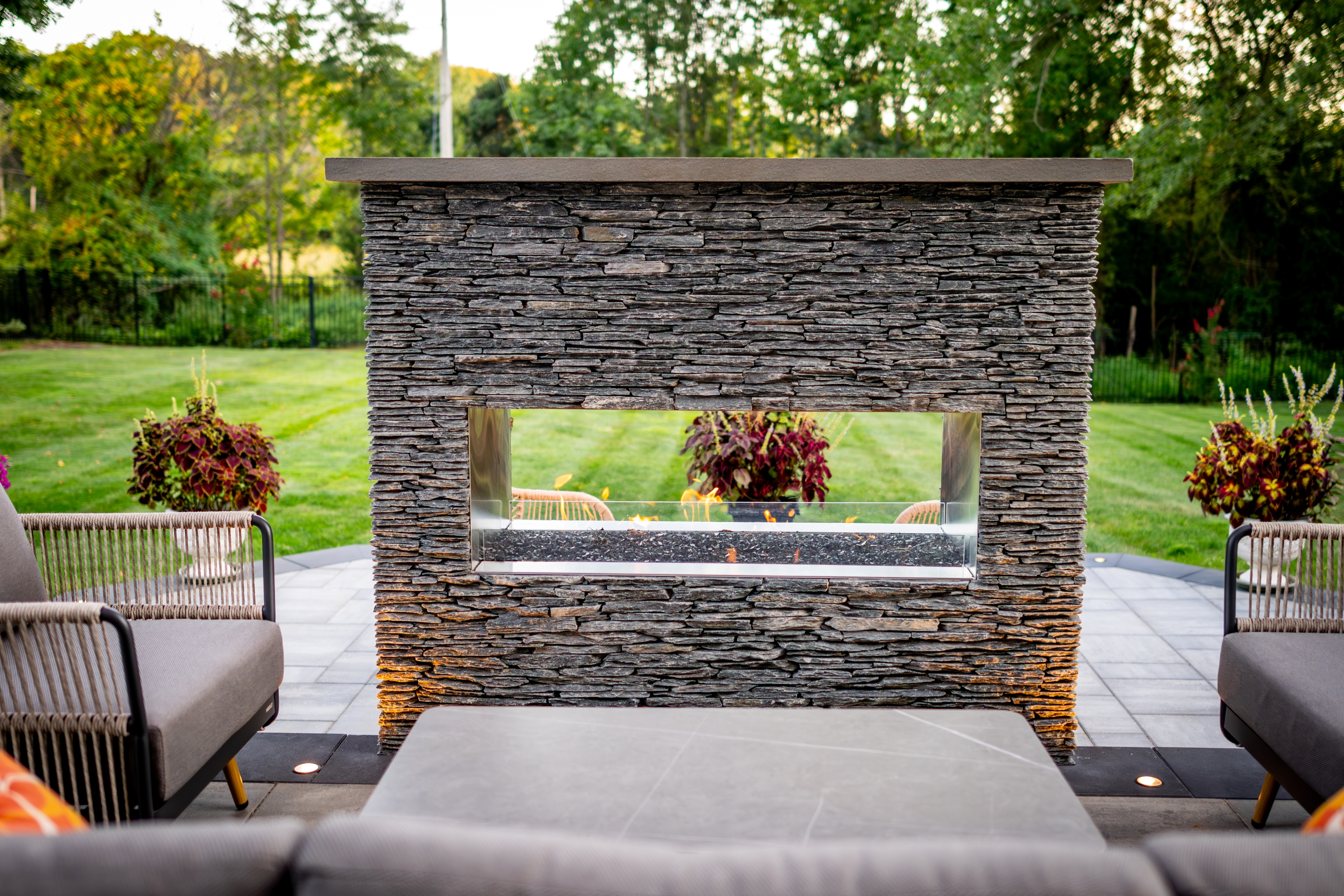 double-sided stone fireplace on raised backyard patio with outdoor seating, overlooking the lawn